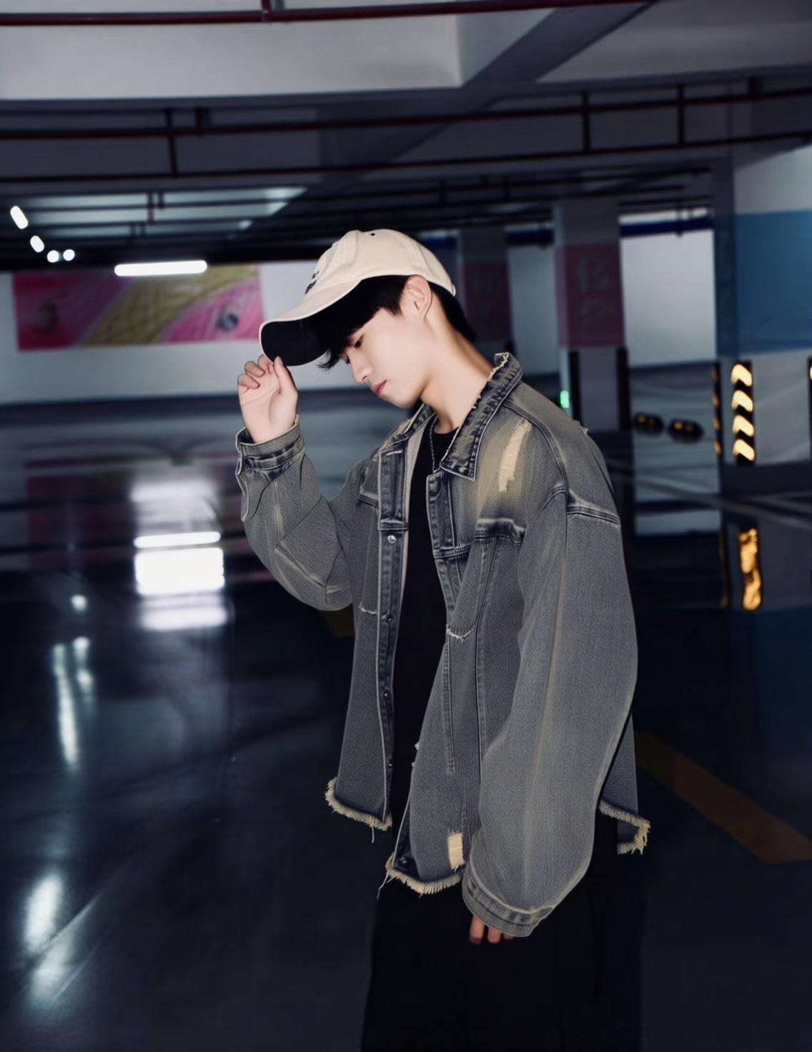 This is a photograph of a young man standing in an empty, dimly lit parking garage. The subject is positioned slightly to the right of the center, facing left with his head slightly tilted downward. He is wearing a light beige baseball cap with a black brim, which he is holding with his right hand as if adjusting it. His hair is dark and partially covers his forehead. He is dressed in a worn, gray denim jacket with visible fraying at the cuffs and hem, over a black t-shirt. His lower body is clothed in black pants. The jackets texture is slightly rough and faded, adding a casual, slightly rugged look to his outfit.The background consists of a dark, polished concrete floor with a few reflective patches, indicating the surface has been recently cleaned. The walls are white with some visible wear and tear, and there are a few indistinct, colorful banners or posters in the far background, adding a touch of color to the otherwise muted setting. The ceiling features exposed beams and fluorescent lights, casting a soft, diffused light that creates gentle shadows on the subjects face and clothes. The overall mood of the photograph is quiet and contemplative.