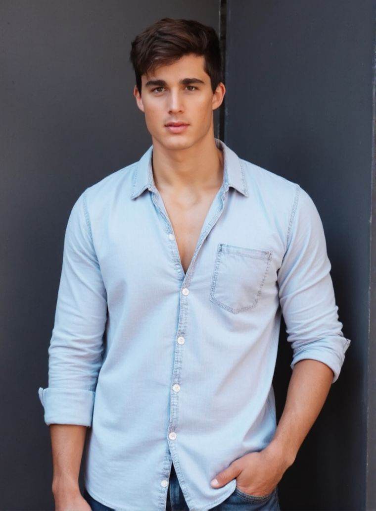 Pietro4Bo4 ,  stands confidently in front of a dark gray wall. He wears a light denim shirt,  unbuttoned at the top on Ten...