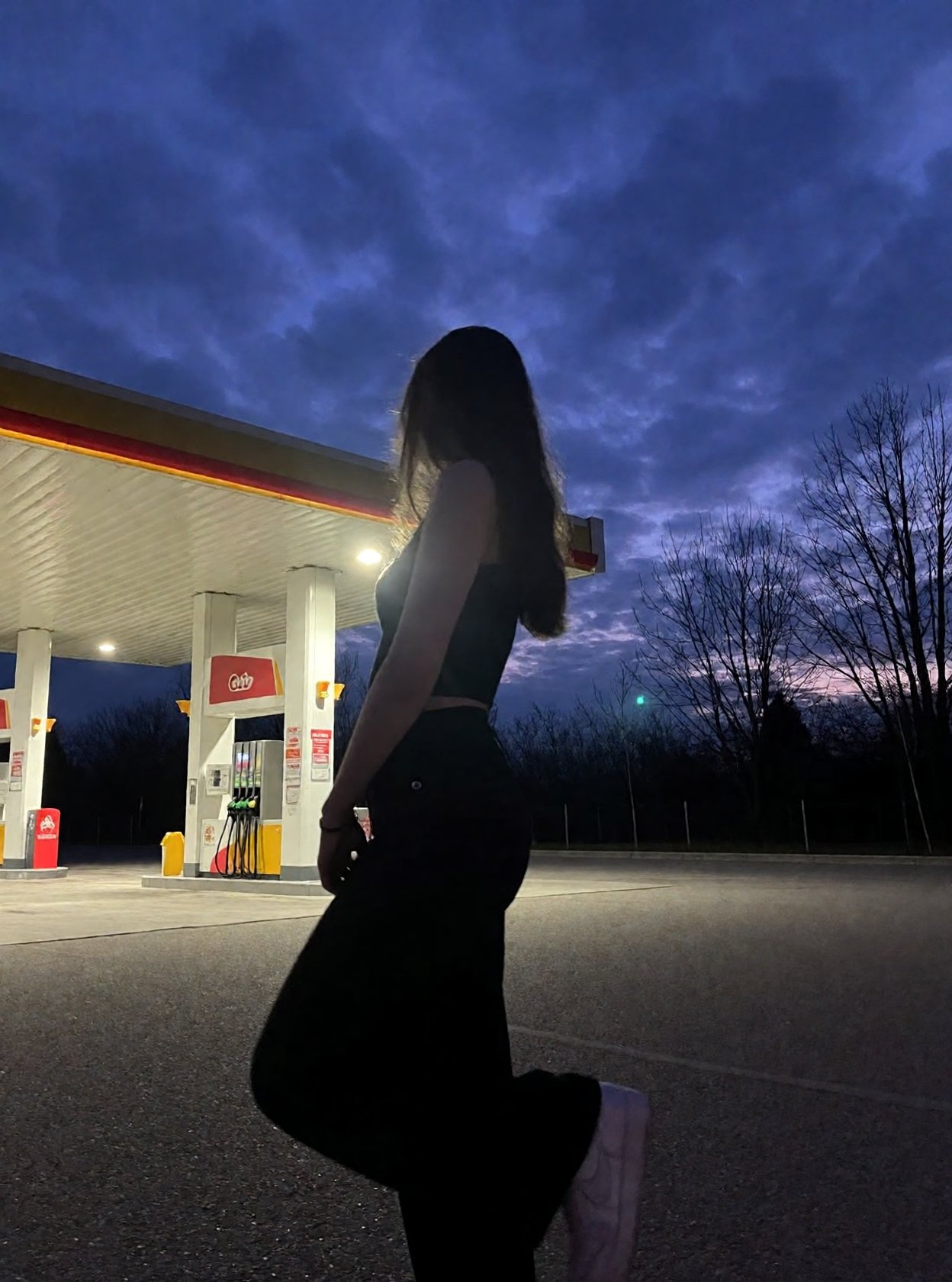 r3ealism,  Captured at dusk,  this low-light photograph features a silhouette of a person standing at a gas station on Ten...