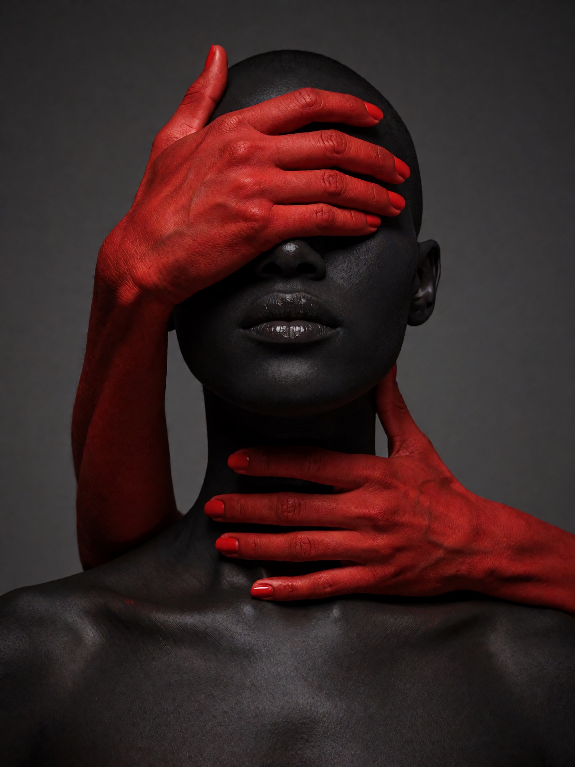 Conceptual portrait of a bald figure with matte black skin against a dark gray backdrop. Two bright red hands emerge from...