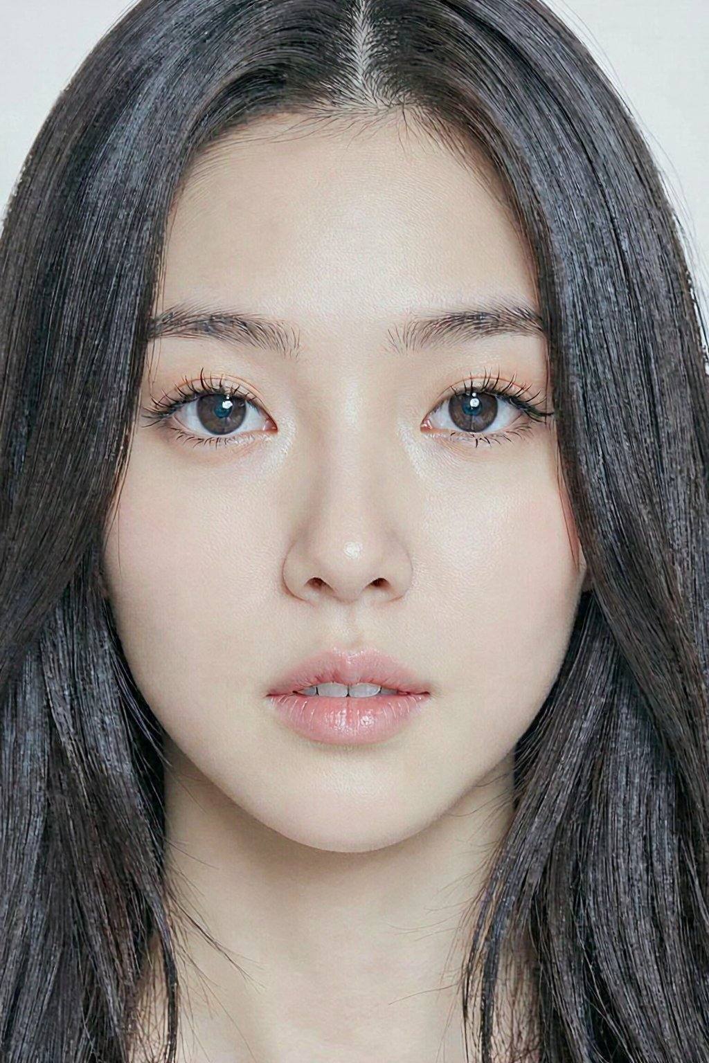 This is a photograph of a young East Asian woman with long, straight black hair framing her face. She has fair skin with a smooth complexion, and her makeup is subtle, highlighting her natural beauty. Her eyes are almond-shaped with dark, well-groomed eyebrows and long, black eyelashes. Her lips are full and slightly parted, showing a natural pink hue. The background is plain white, making her facial features stand out prominently. The lighting is soft, enhancing her clear skin and giving a gentle glow to her complexion.