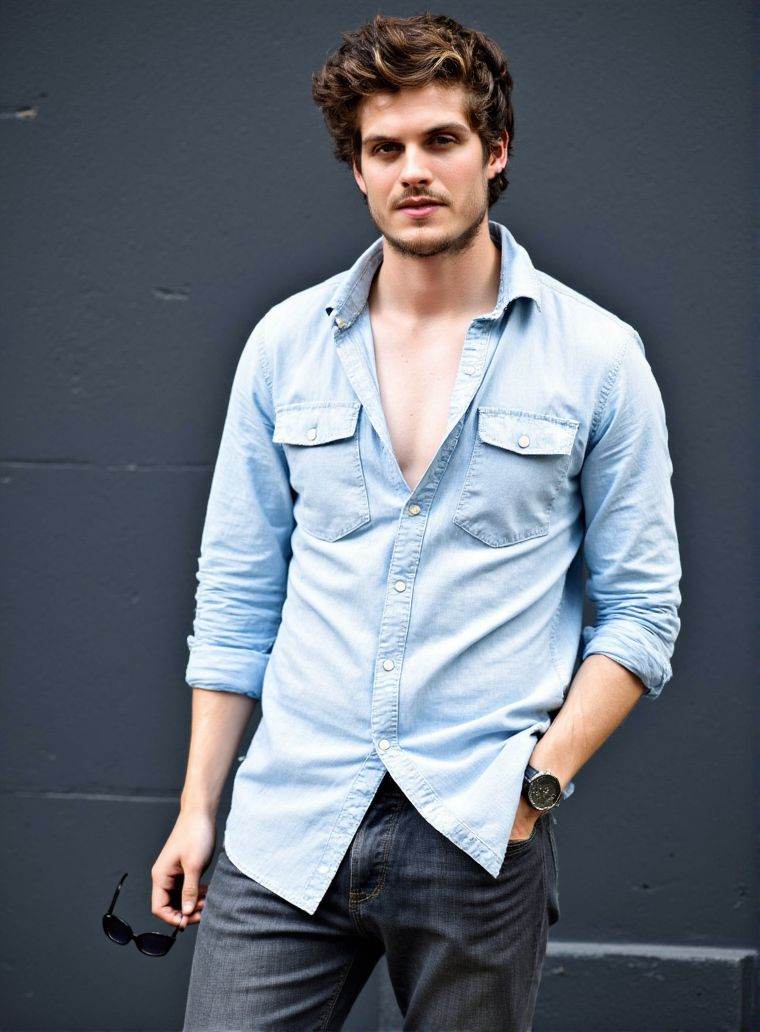 danielsharman ,  stands confidently in front of a dark gray wall. He wears a light denim shirt,  unbuttoned at the top on...