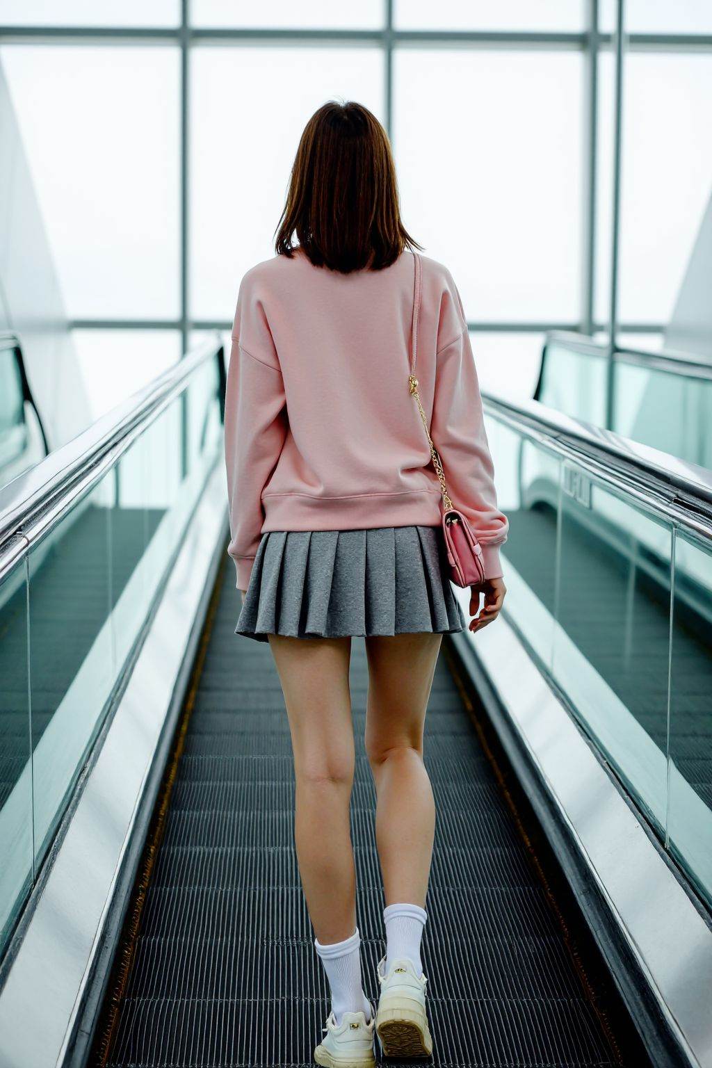 This is a photograph taken in a modern, well-lit indoor space, likely a shopping mall or airport, featuring a young woman ascending a moving walkway. The woman is standing with her back to the camera, facing the escalator, which is positioned centrally in the image. She has a fair complexion and shoulder-length brown hair. She is wearing a pastel pink sweatshirt and a short, pleated grey skirt that reveals her thighs. Her outfit is complemented by white ankle socks and white sneakers with black soles. She also carries a small pink shoulder bag on her right side.The background shows large glass windows that let in natural light, creating a bright and airy atmosphere. The escalator is flanked by sleek, glass walls, and the walkway has a black rubber surface. The image is taken from a low angle, emphasizing the woman's height and giving a sense of perspective. The overall scene is clean and modern, with a focus on the woman's attire and the architectural elements of the space. The photograph captures a moment of casual, everyday life in a public setting.