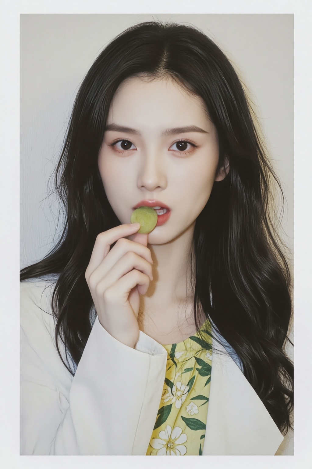 ZOZ_PLD,a photo-realistic portrait of a young woman with long,wavy black hair,holding a small,green fruit in her mouth,she is positioned in the middle of the frame,looking directly at the viewer with a neutral expression,her eyes are a striking brown color,and her lips are slightly parted,revealing a hint of her teeth,she has a slim body and is wearing a white blouse with a floral pattern,the background is a plain,light-colored wall,and the image has a soft,ethereal quality to it,the woman is holding the fruit with one hand and her fingers are delicately placed near her mouth,as if savoring the moment,the image is framed with a white border,giving it a clean and professional look,