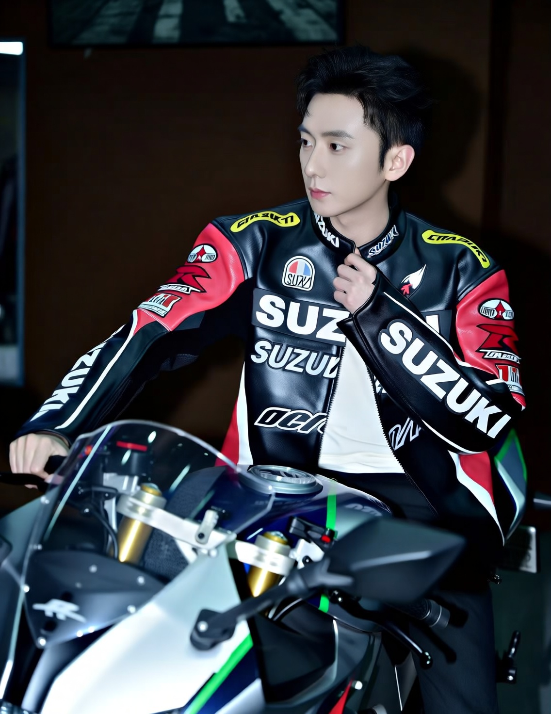 This is a photograph of an Asian man with short, dark hair, sitting on a sleek, modern motorcycle. He is wearing a black and red racing jacket adorned with various sponsor logos, including SUZUKI prominently displayed in white on the chest and sleeves. The jacket has a glossy texture with a mix of matte and reflective surfaces, and it is zipped up slightly to reveal a white t-shirt underneath. The mans right hand is resting on the handlebar of the motorcycle, while his left hand is lightly gripping the collar of his jacket. The motorcycle is predominantly black with green accents, featuring a clear windshield and a sleek, aerodynamic design. The background is dimly lit, with a blurred, dark brown wall and some indistinct objects, suggesting an indoor setting, possibly a garage or workshop. The mans expression is neutral, and he is looking slightly to the left of the camera. The lighting in the photograph is soft, highlighting the mans face and the motorcycle, while the background remains in shadow, creating a contrast that draws attention to the subject. The overall style of the photograph is modern and professional, emphasizing the subjects association with motor sports.