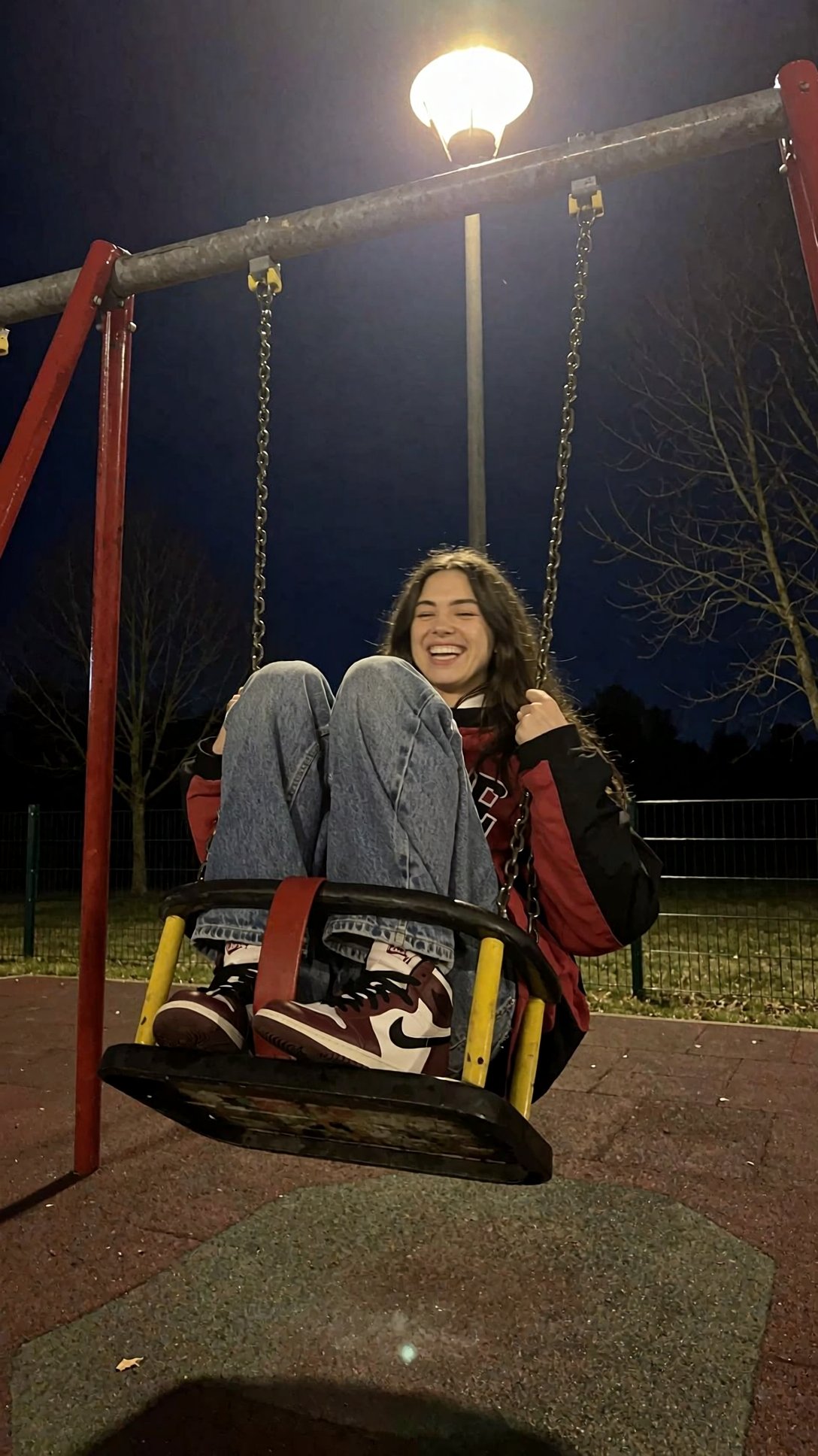 r3ealism,  A woman with long dark hair sits laughing on a child-sized park swing at night,  their legs tucked up against t...