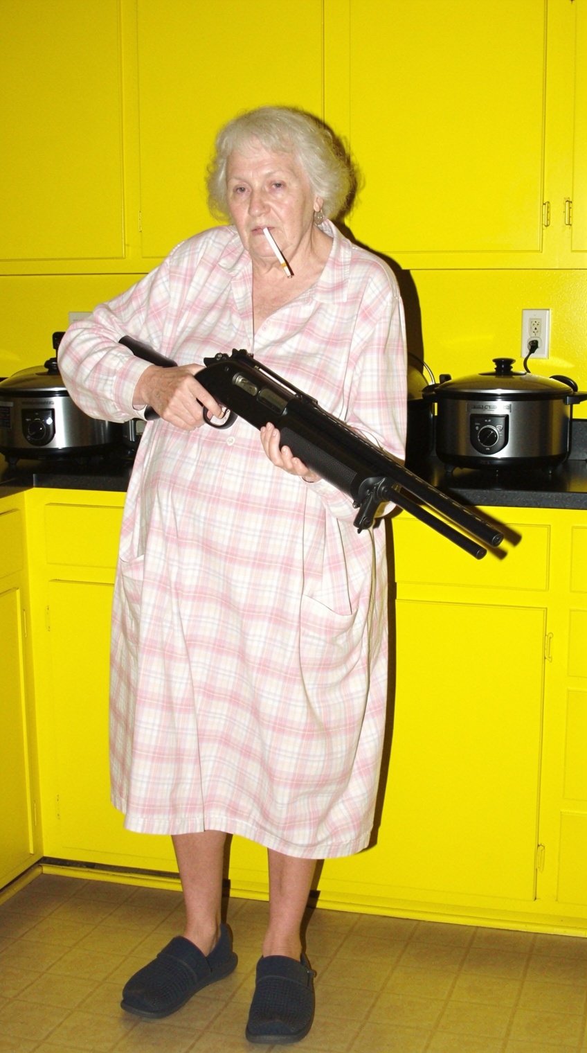 A full-body shot of an elderly woman standing in a domestic kitchen with bright yellow walls. She is wearing a loose,  lig...