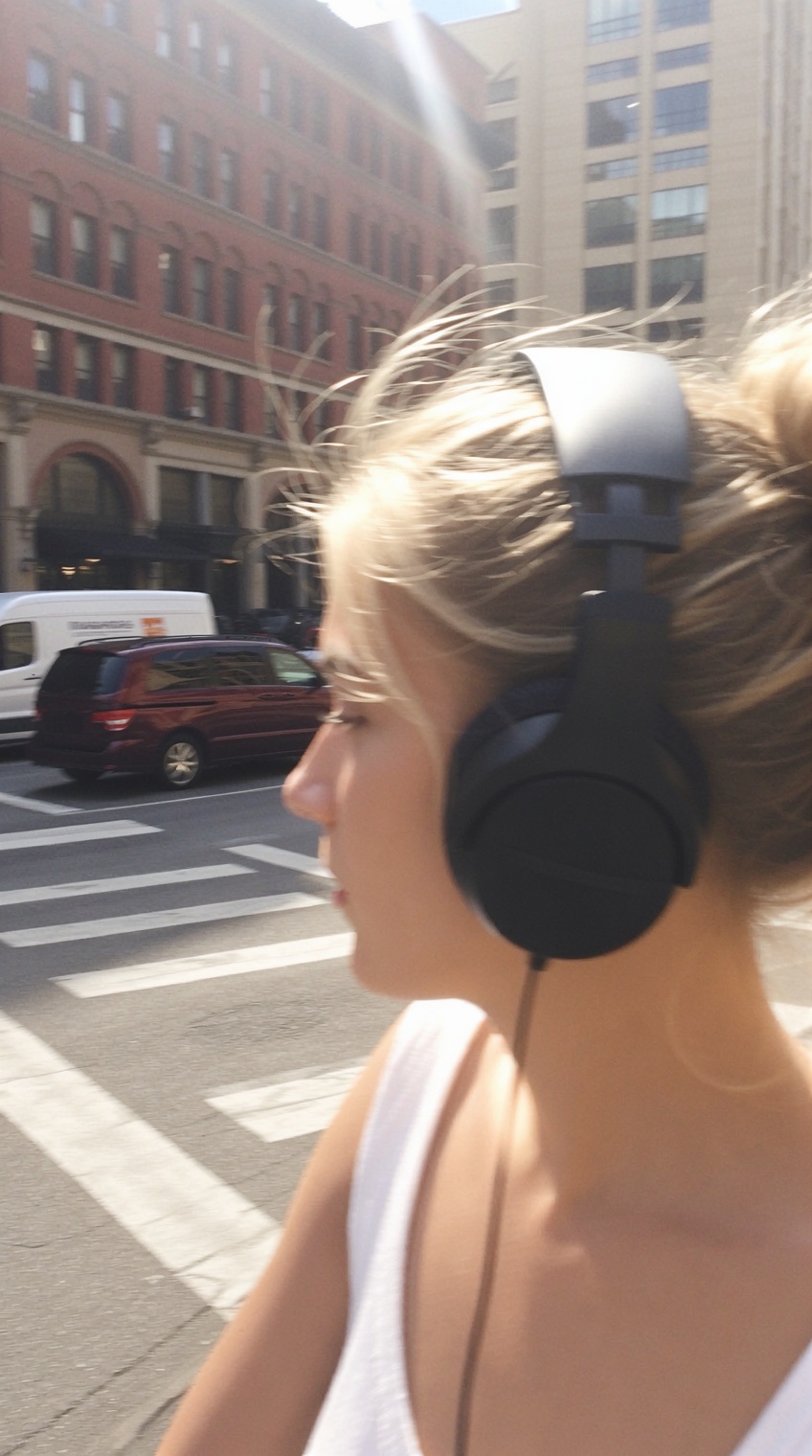 a young woman with light,  windswept hair tied in a messy bun walks across a sunlit city street,  wearing large black over...