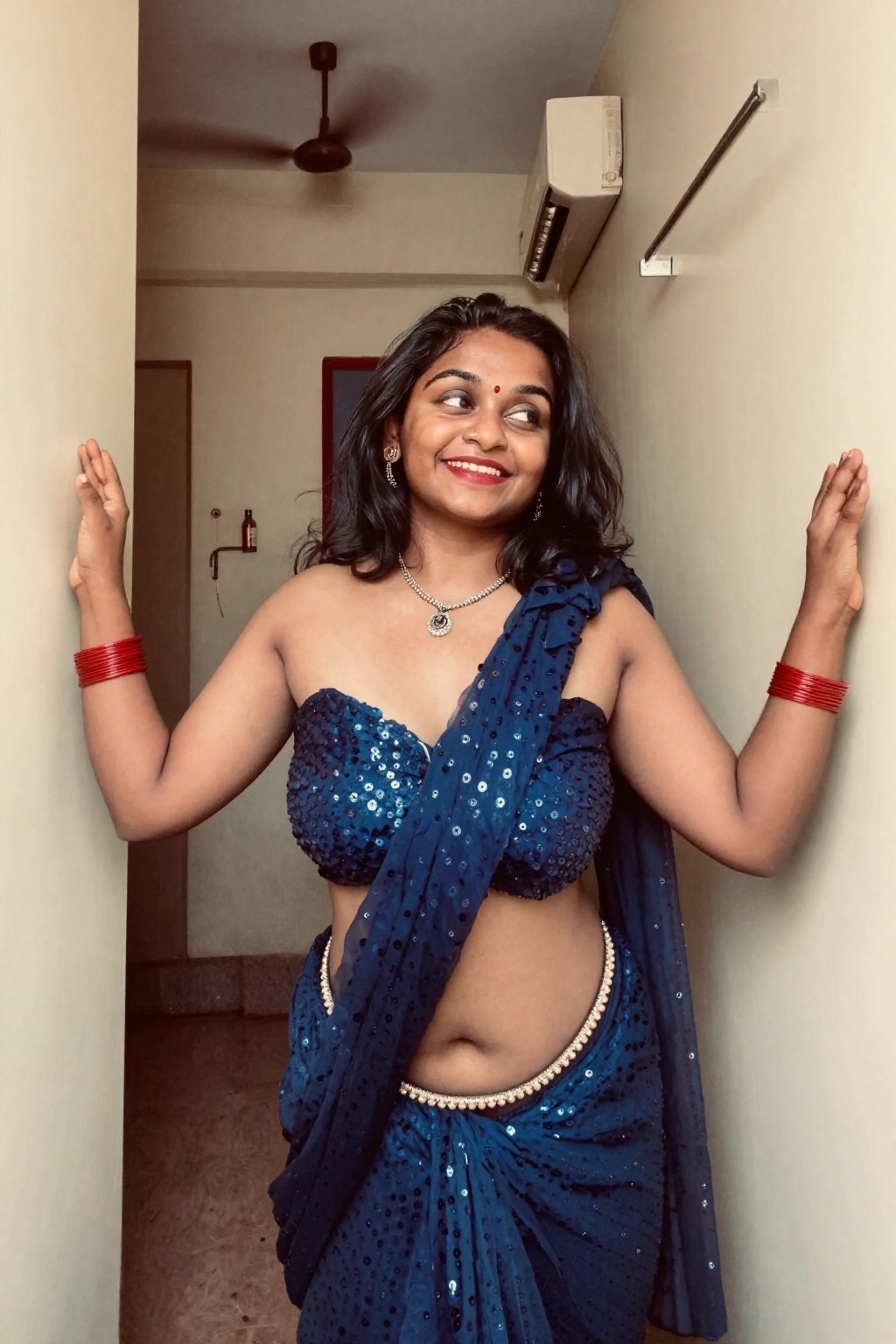 Photograph of a smiling Indian woman standing in a narrow hallway. She has olive skin black hip-length hair and is wearing...