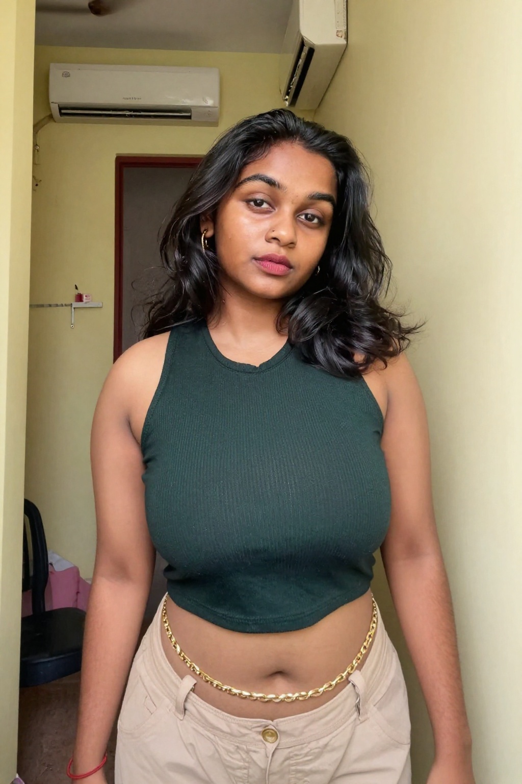 Photograph of a young South Asian woman with medium brown skin and long wavy black hair. She stands in a narrow hallway wi...