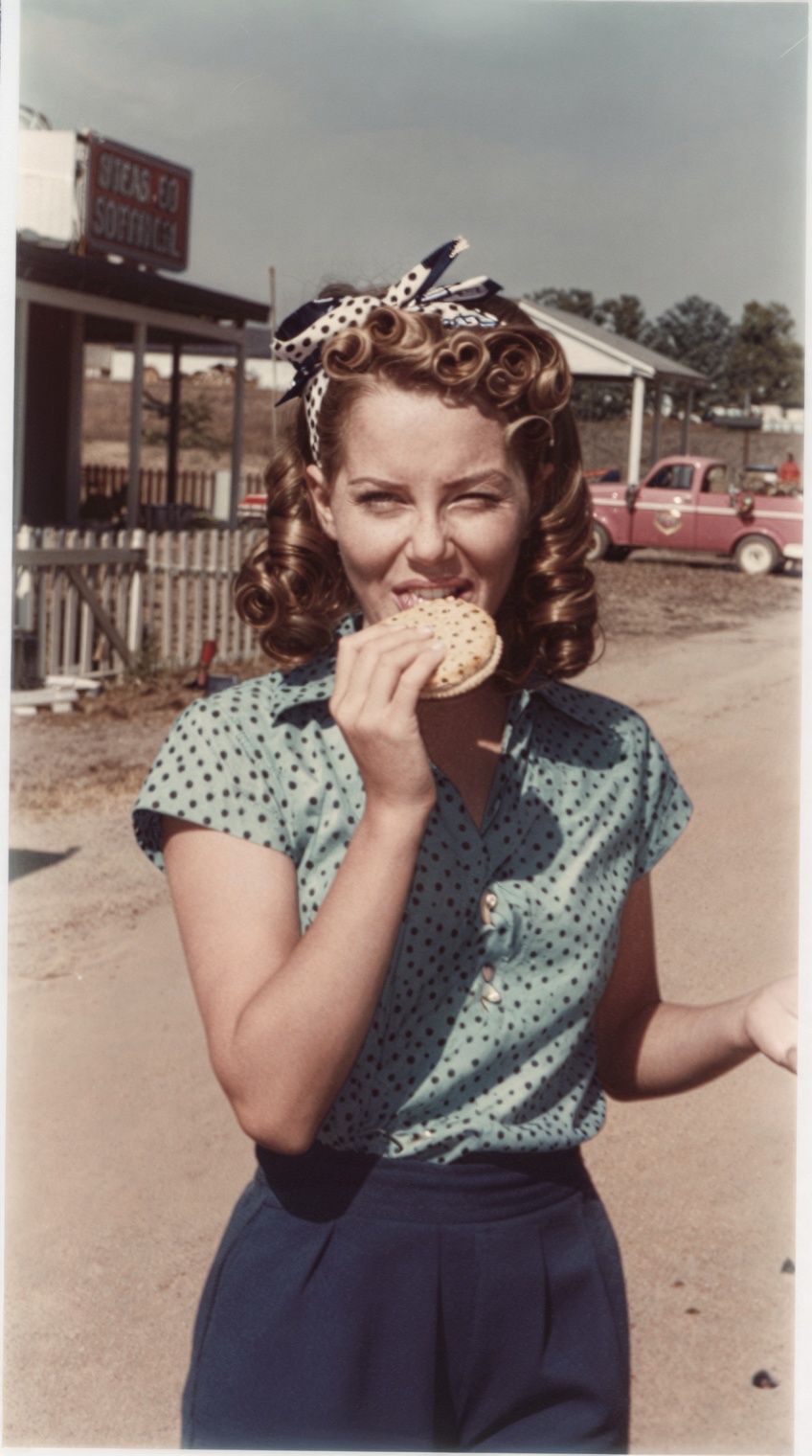 A candid vintage photo from the 1960s showing a 20-year-old girl in a rural American town. She’s caught mid-bite while eat...