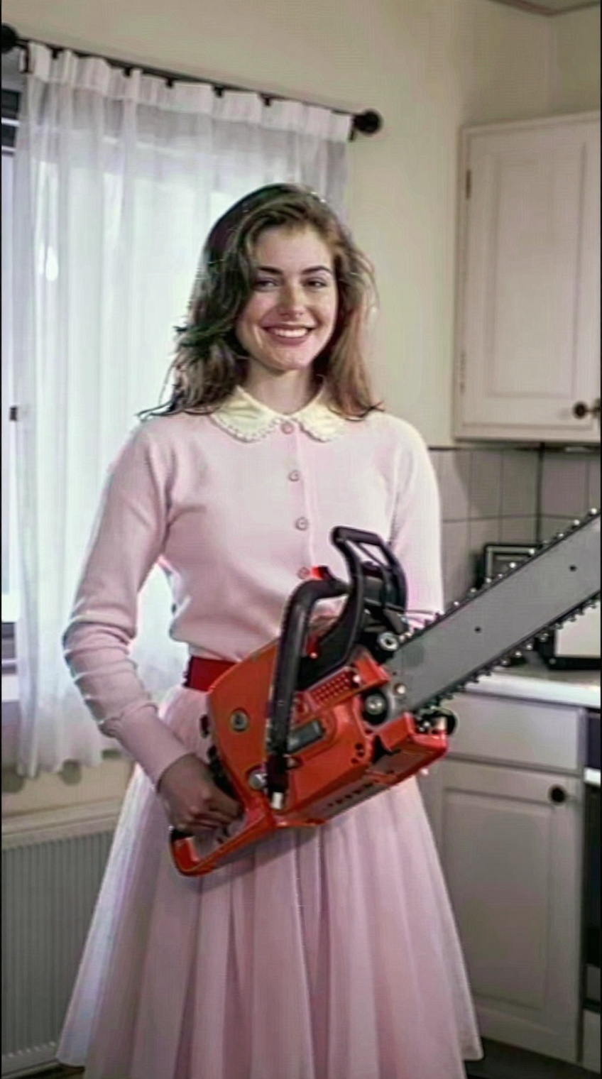 A smiling young woman with light brown hair stands indoors,  holding a large red and silver chainsaw horizontally in front...