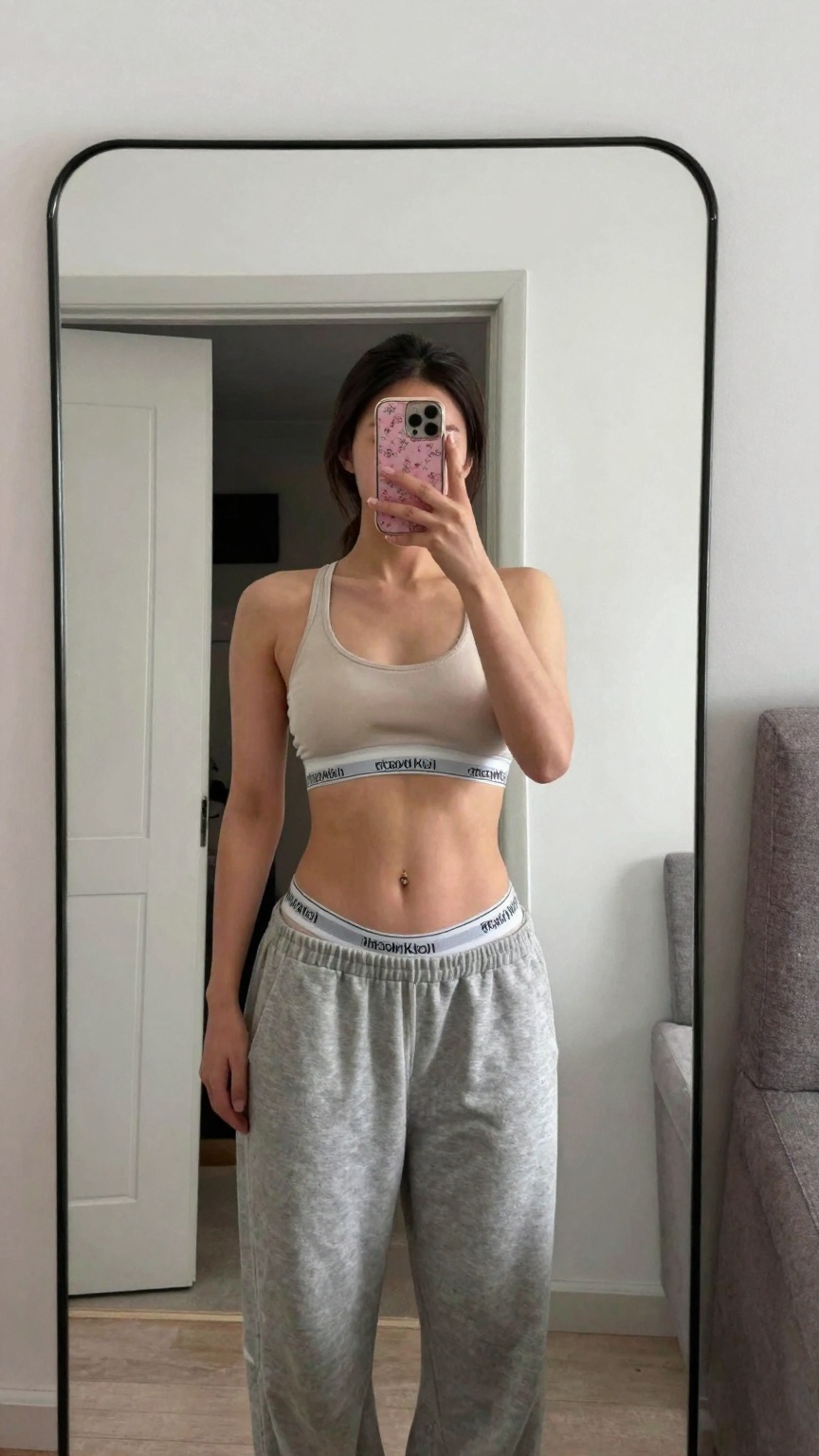 Photograph of a young woman taking a mirror selfie in a minimalist room. She has light skin dark straight hair tied back a...