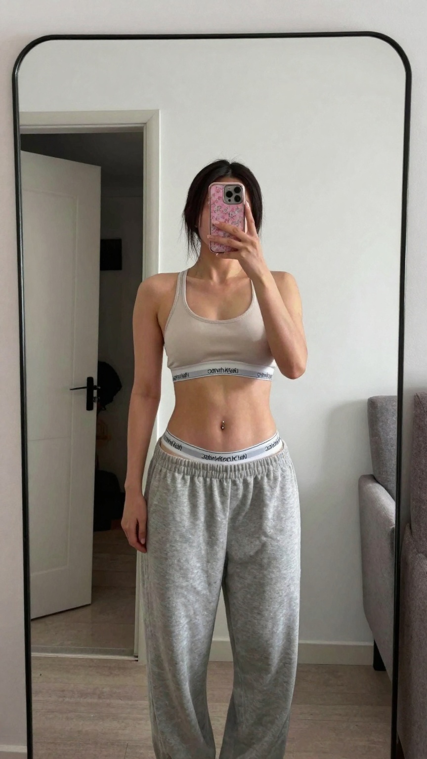 Photograph of a young woman taking a mirror selfie in a minimalist room. She has light skin dark straight hair tied back a...