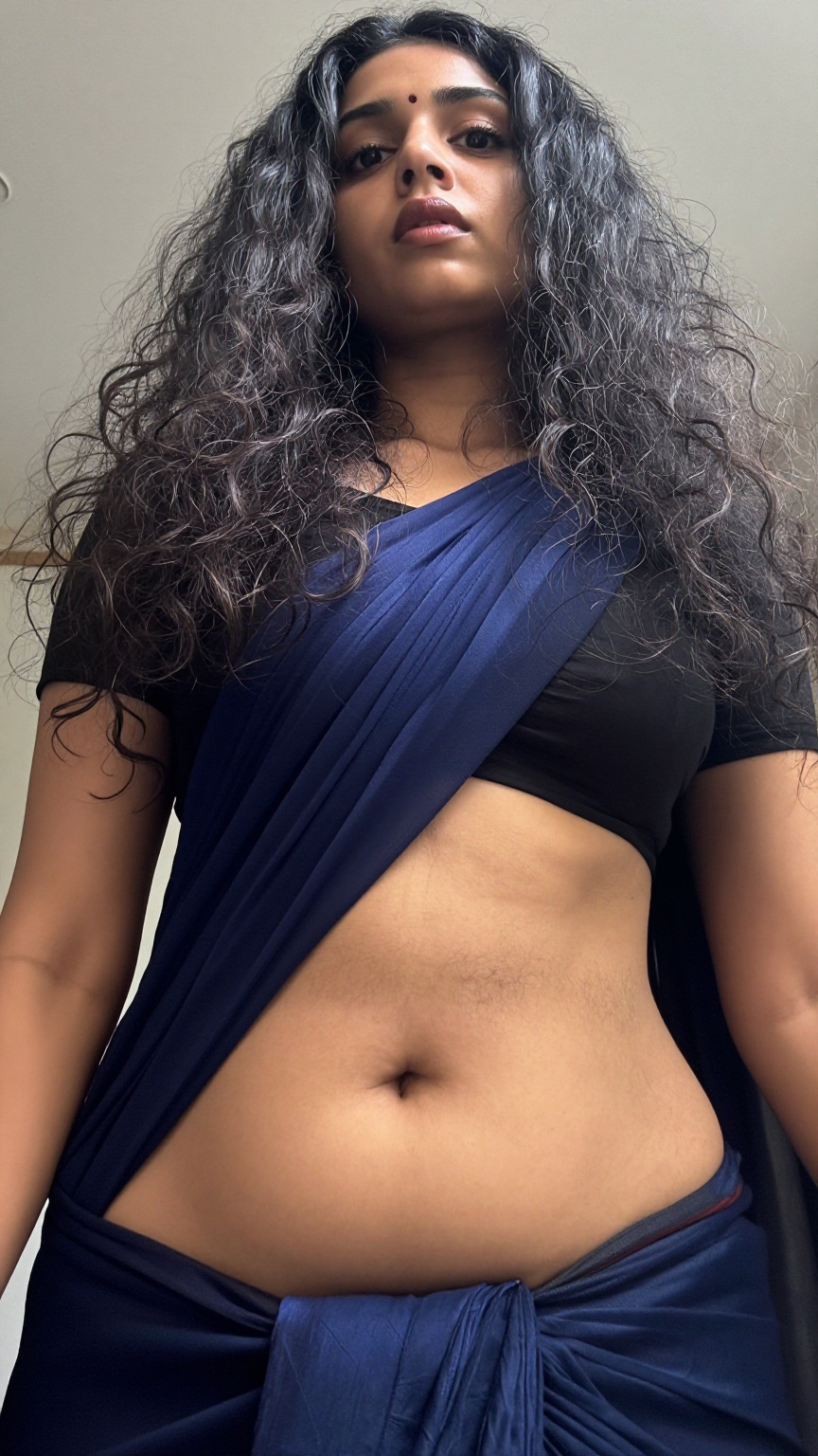 A worm’s-eye view low-angle shot captures a Mallu young slim woman with a seductive expression. Her long,  wavy hair casca...