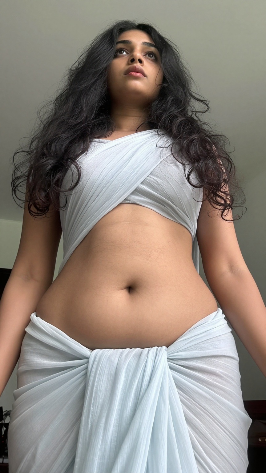A worm’s-eye view low-angle shot captures a Mallu young slim woman with a seductive expression. Her long,  wavy hair casca...
