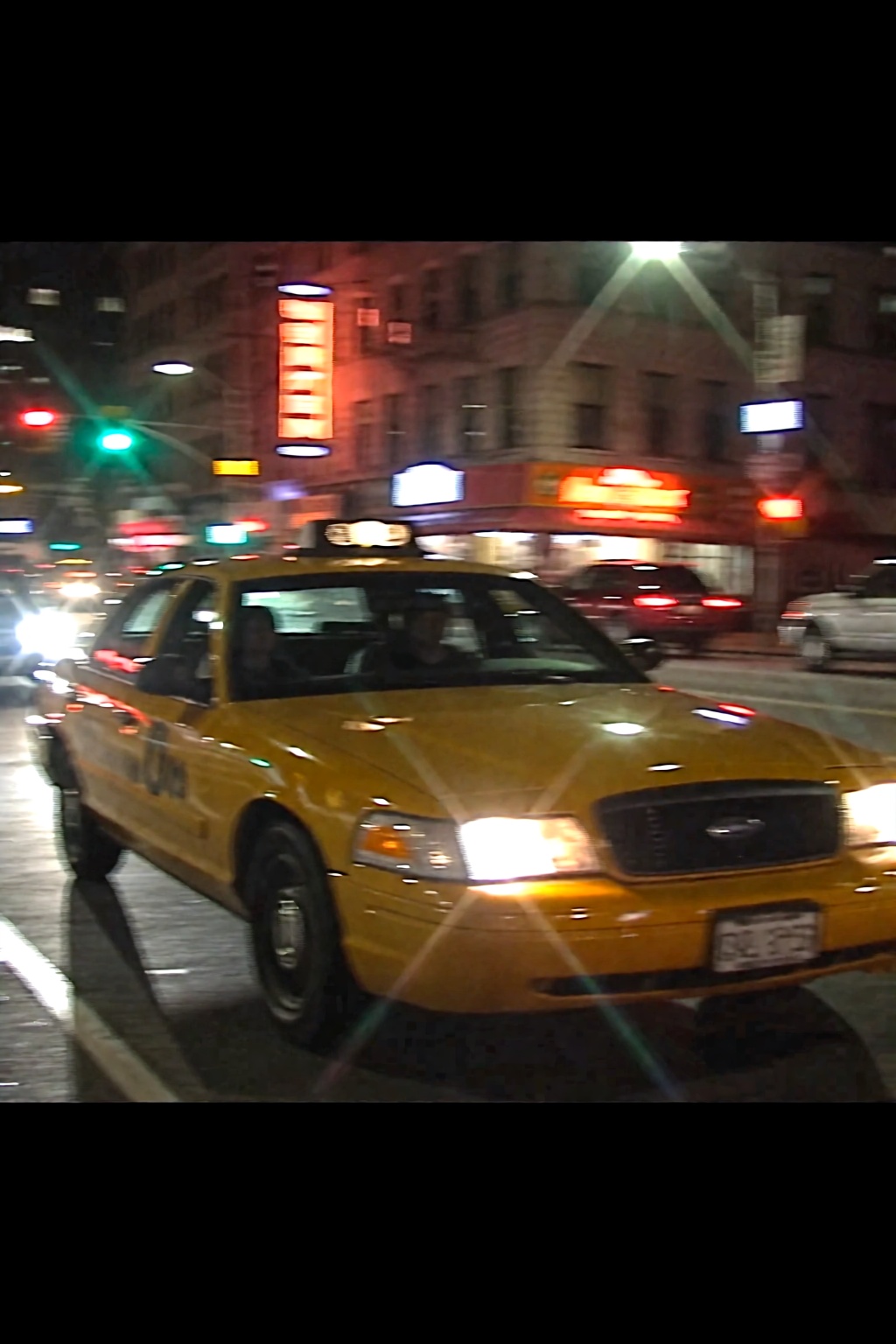 A camcorder video still capturing a yellow Ford Crown Victoria taxi speeding through a city at night,  shown in a blurry,...