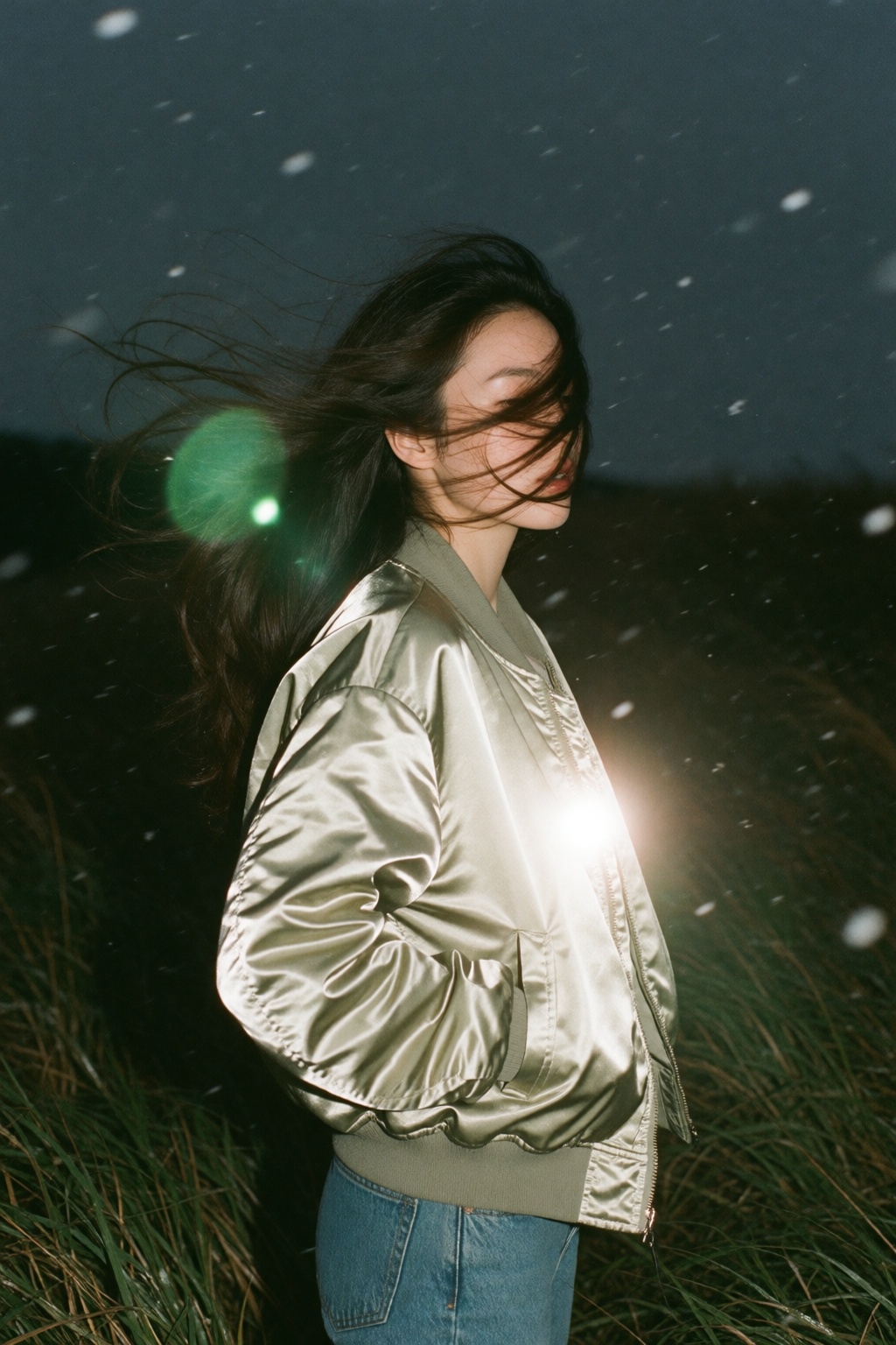 An experimental analog film snapshot of a young woman standing in a grassy field at night under a dark twilight sky. Captu...