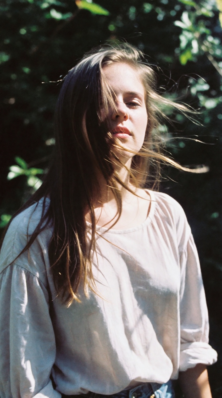 A candid,  atmospheric medium shot outdoor portrait of a young woman with long,  flowing hair blowing across her face. She...