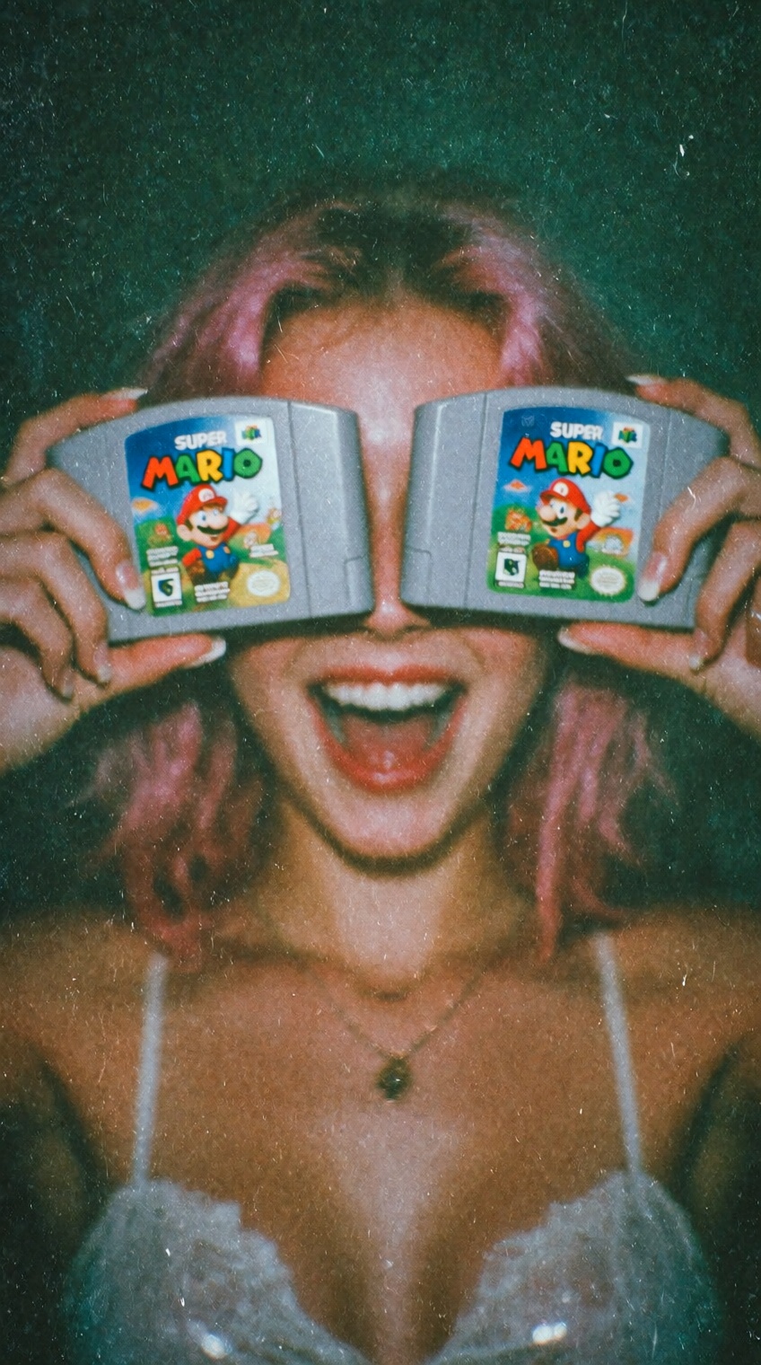 A playful,  nostalgic photo of a young woman with pink-tinged hair holding two N64 super mario game cartridges over her ey...