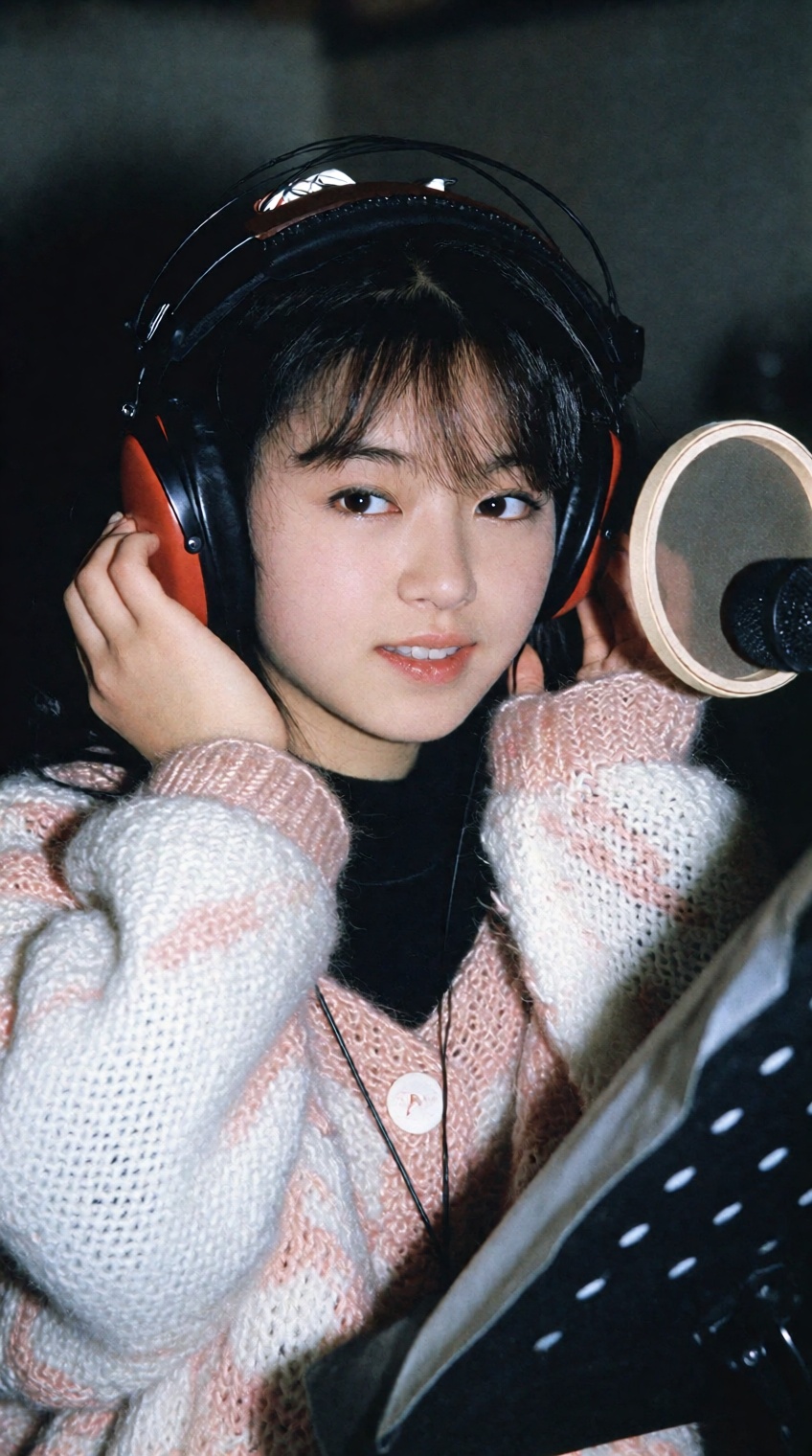 A young woman with short dark hair and bangs sits in a dimly lit recording studio,  wearing large over-ear headphones with...