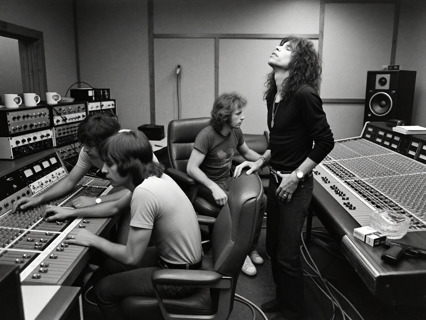 {"high_level_description":"A gritty,  high-contrast monochrome 1970s analog recording studio scene. Three men are focused...