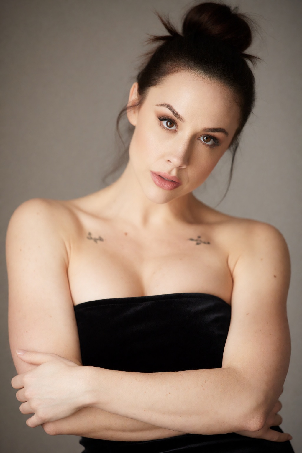 An artistic beauty portrait showing woCh40n with dark hair in a messy,  chic topknot,  crossing her arms loosely in front...