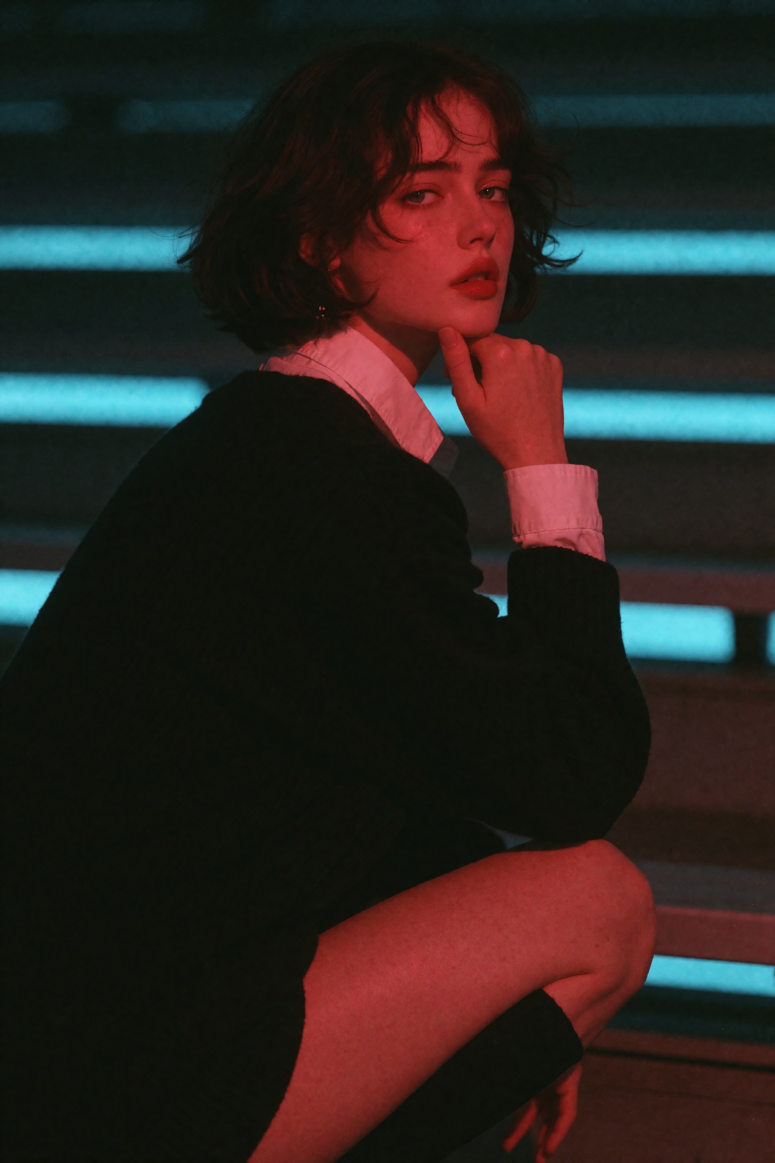 A young woman with a dark bob crouches on slatted bleachers,  glancing over her shoulder,  resting her chin on her hand. S...