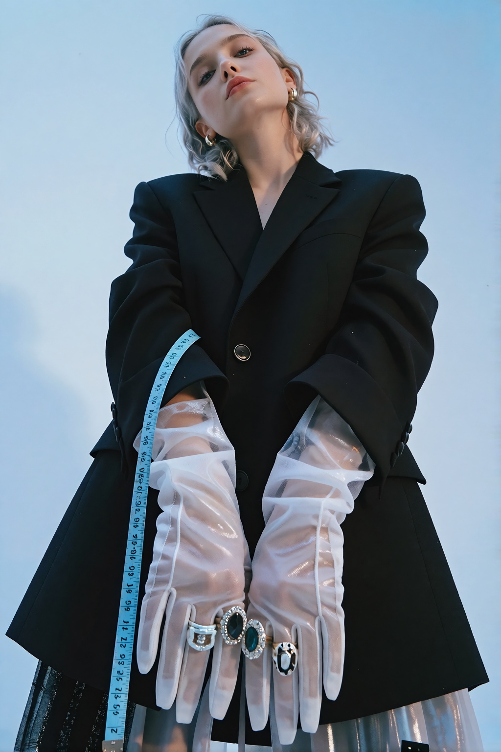 A low-angle fashion portrait of a silver-haired person in a black blazer against a solid pale blue backdrop. They wear dra...