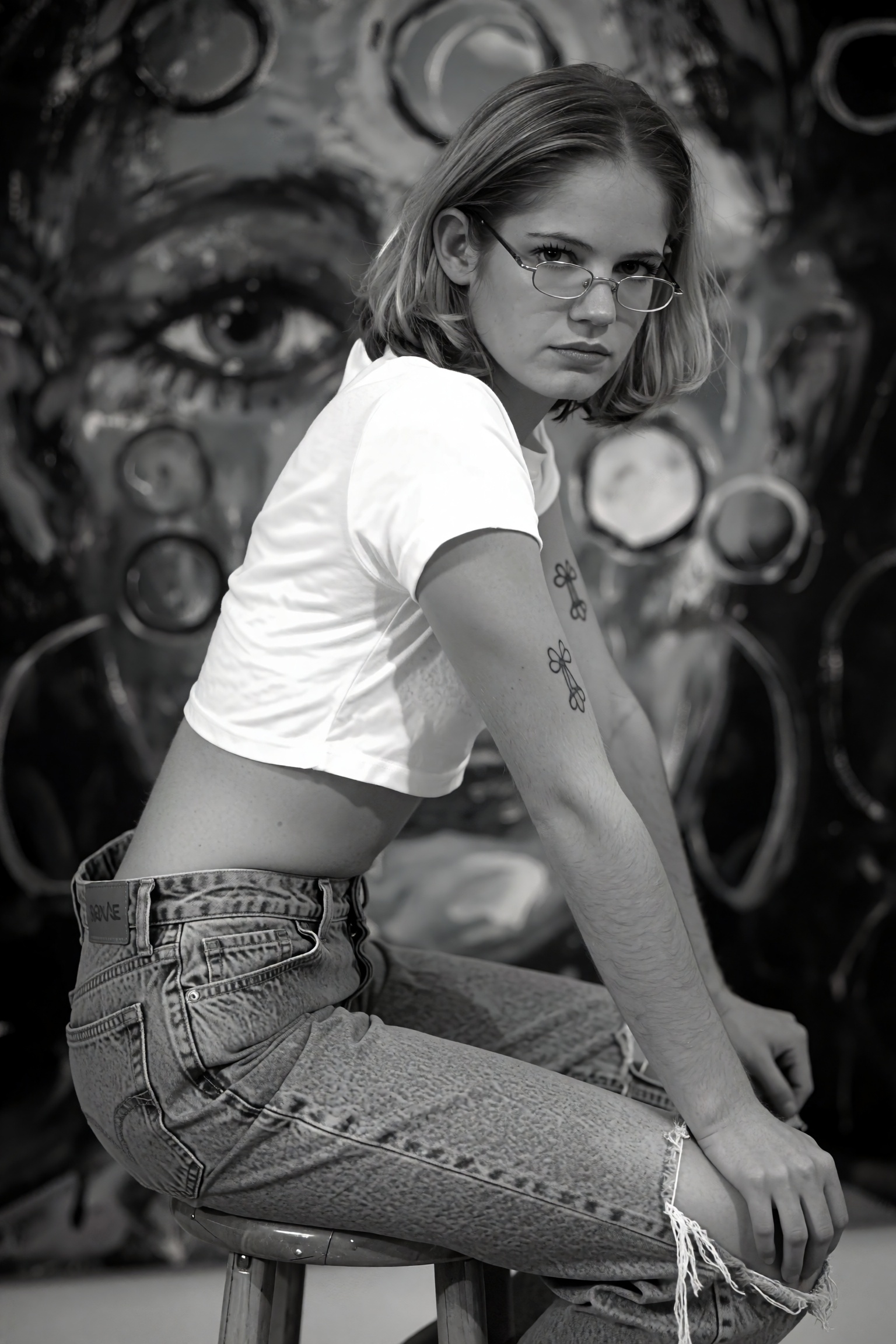 A black and white photograph of a ***** woman sitting on a stool in the middle of the frame,  wearing a cropped white crop...