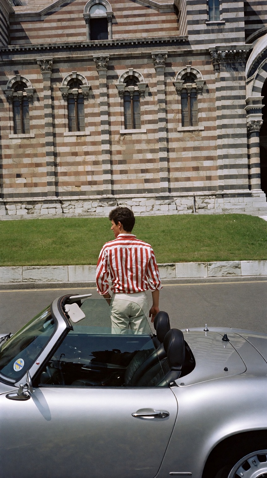@charleshtraub,  A man in a red-and-white striped shirt stands behind a silver convertible. He looks toward a massive buil...