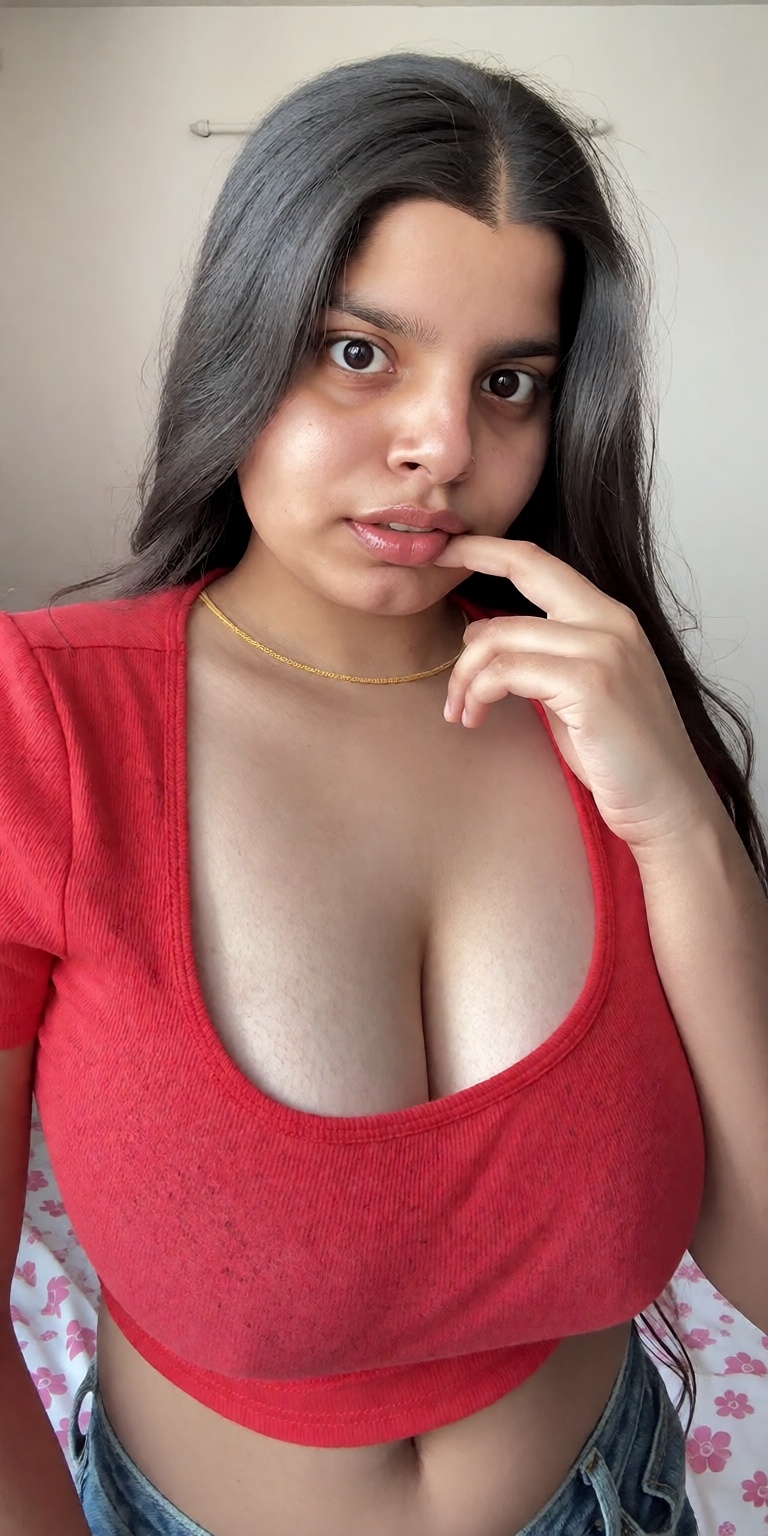 A young Indian woman with long silky black hair and a small bindi sits casually indoors,  her large heavy sagging breasts...