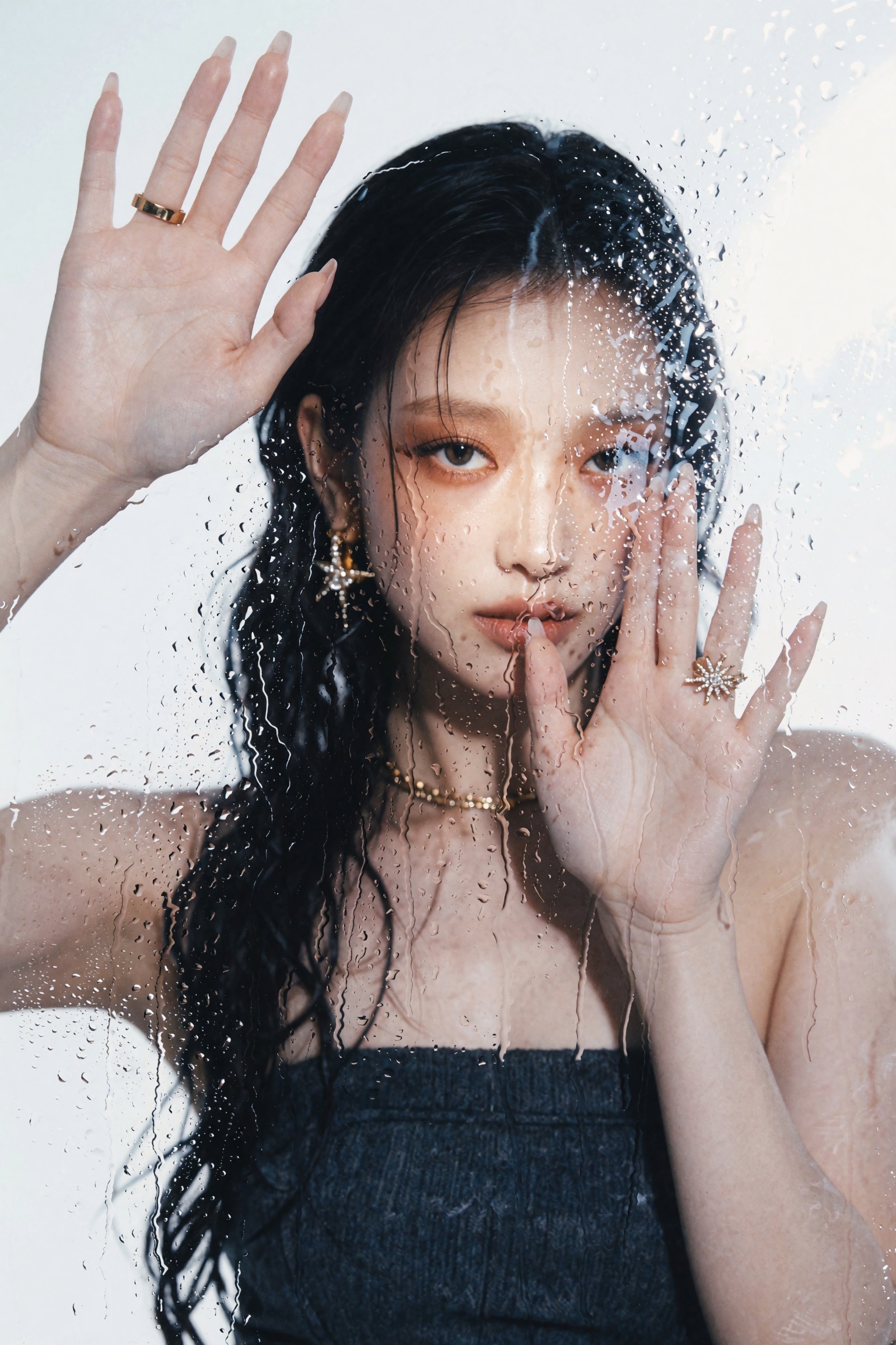 A young woman with wet dark hair clinging to her face and neck presses both palms against a transparent glass pane covered...
