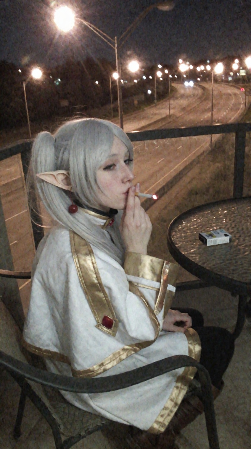 A candid nighttime snapshot of an elf mage cosplaying Frieren sitting on an apartment balcony chair. She has silver-white...