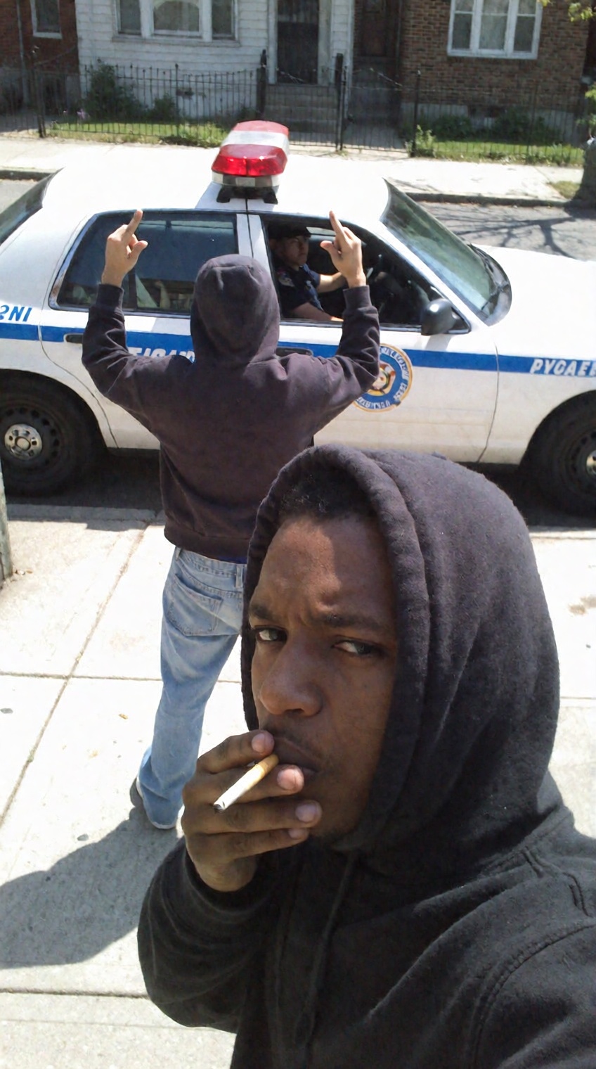A candid urban street selfie captured on a sunny residential sidewalk. In the immediate lower foreground,  a young Black m...