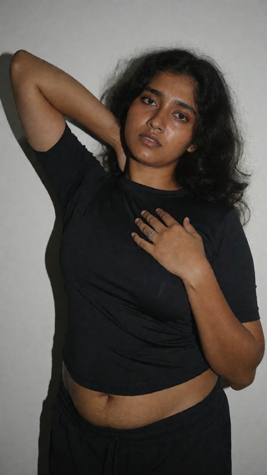 Photograph of a young South Asian woman with medium brown skin and long,  wavy black hair. She is standing against a plain...