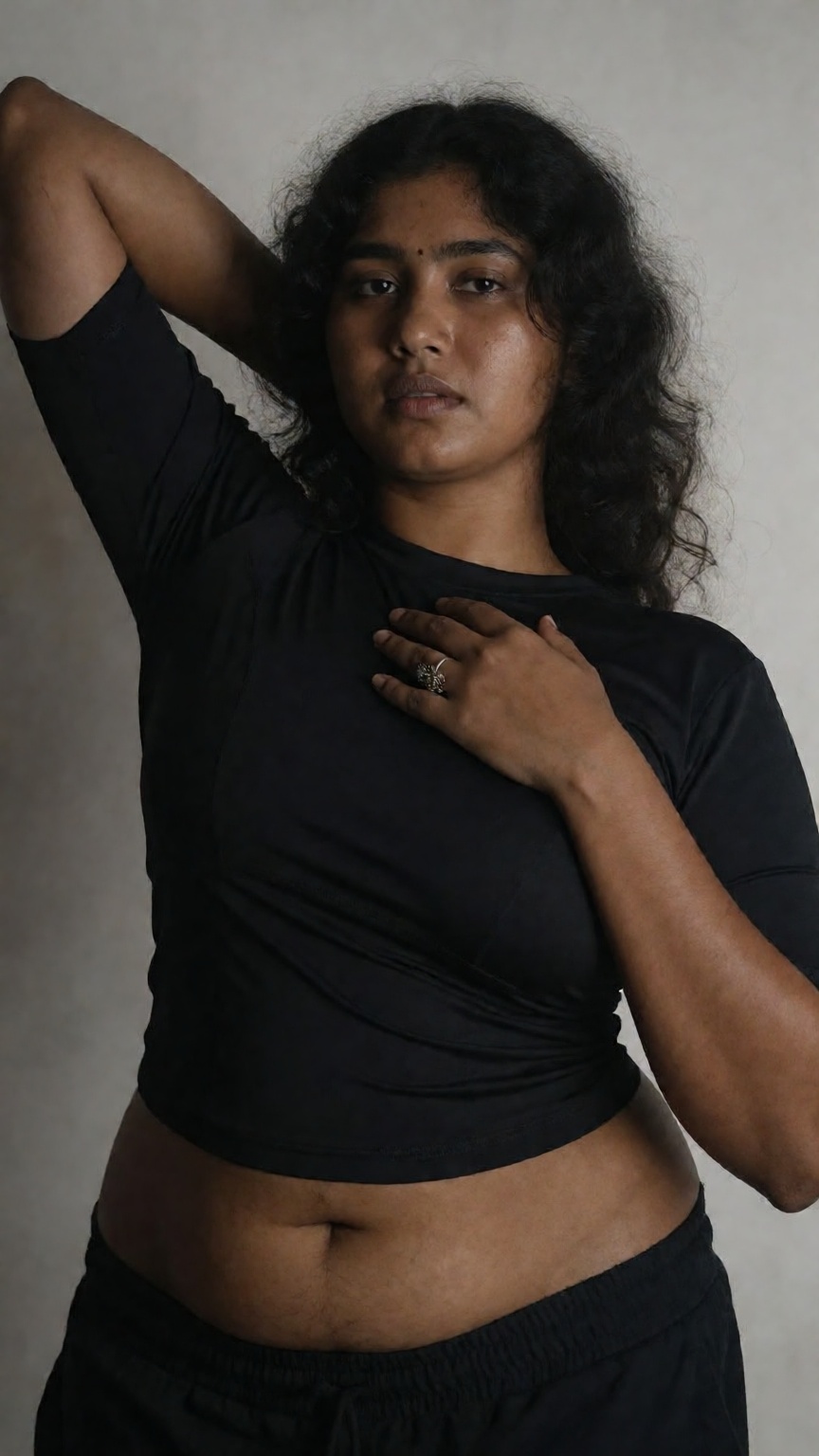 Photograph of a young South Asian woman with medium brown skin and long,  wavy black hair. She is standing against a plain...