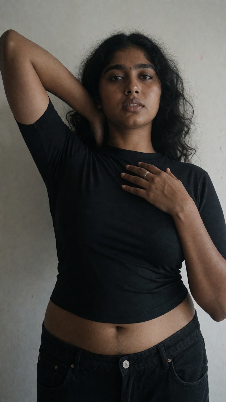 Photograph of a young South Asian woman with medium brown skin and long,  wavy black hair. She is standing against a plain...