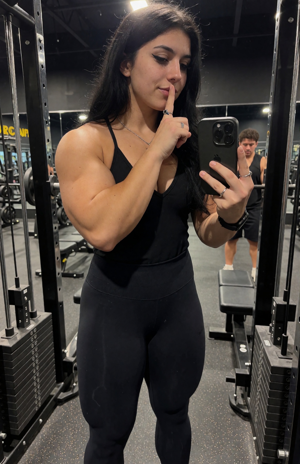 Your favorite goth muscle mommy stands between two cable stations in a modern commercial gym,  caught in candid phone phot...
