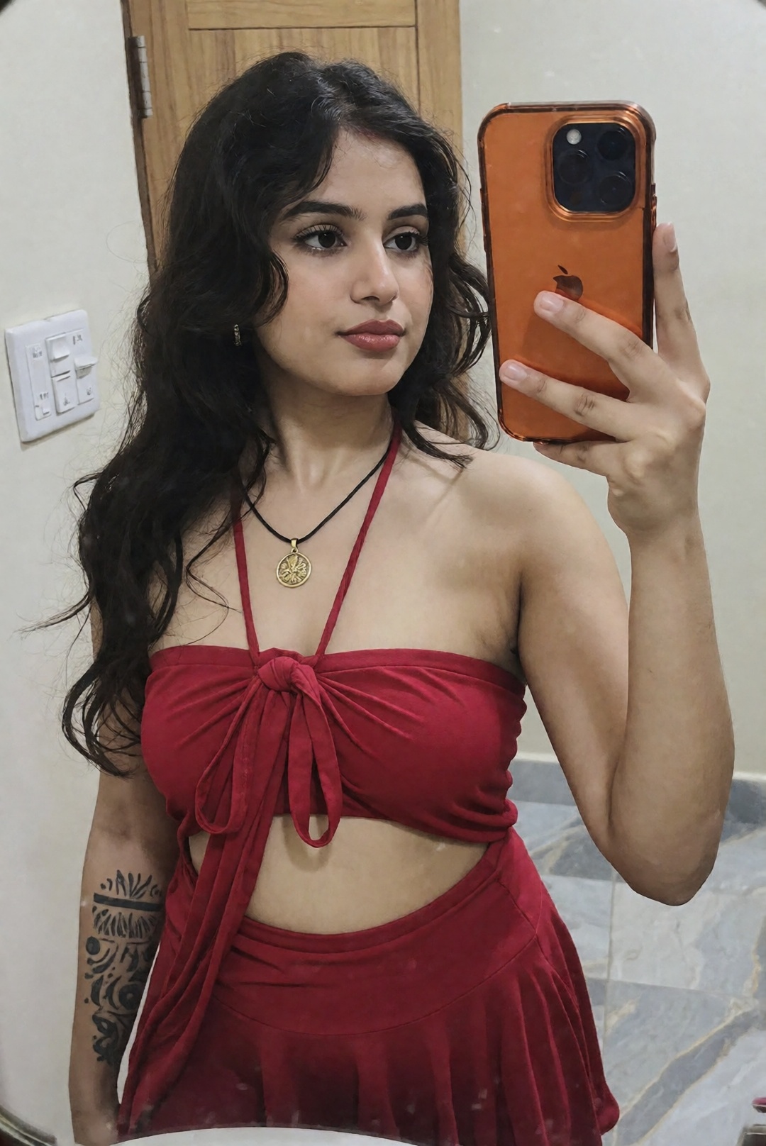 The image shows a south asian indian woman taking a mirror selfie. The individual is wearing a red outfit that includes a...