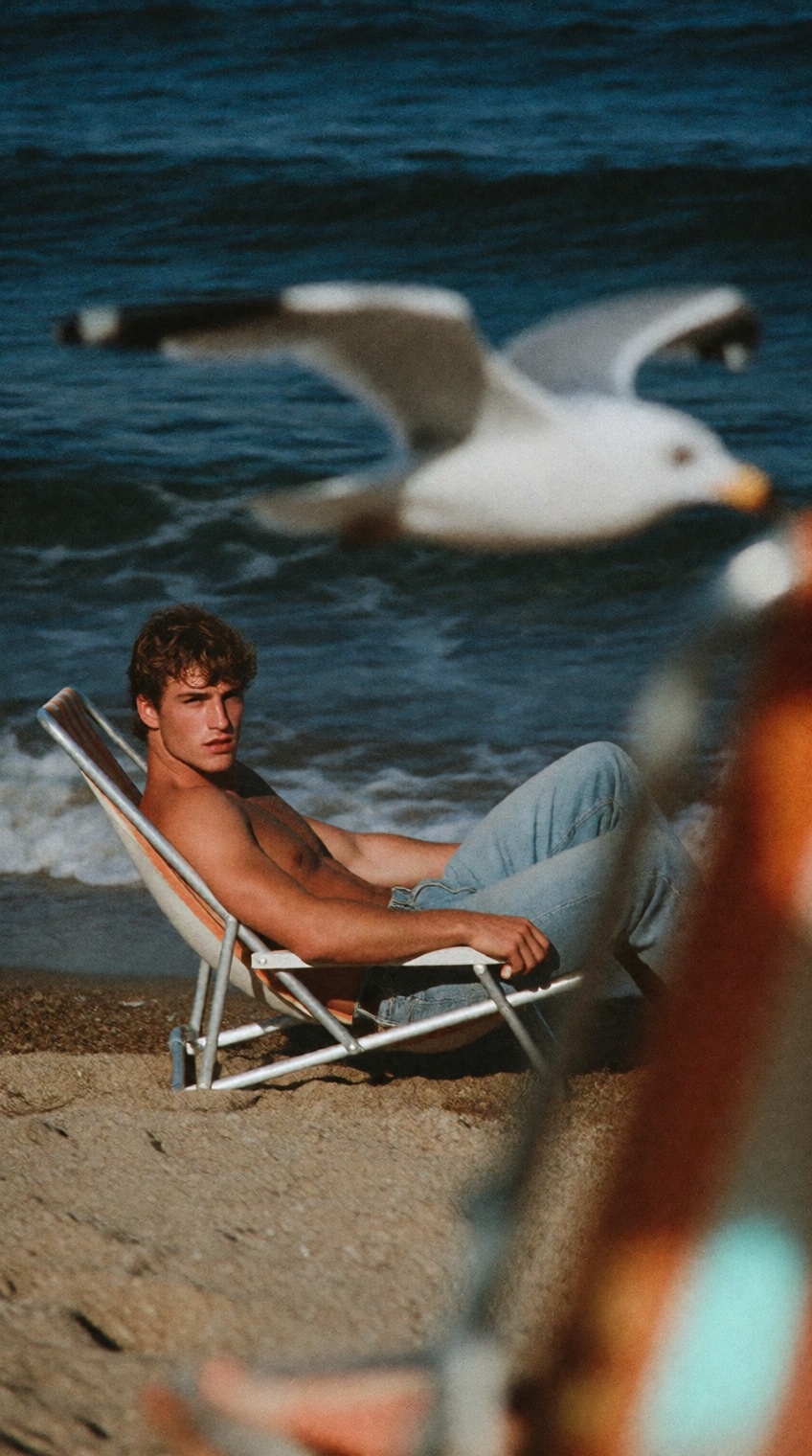 a shirtless man with curly brown hair reclines in a folding beach chair on tan sand,  wearing light blue jeans and gazing...