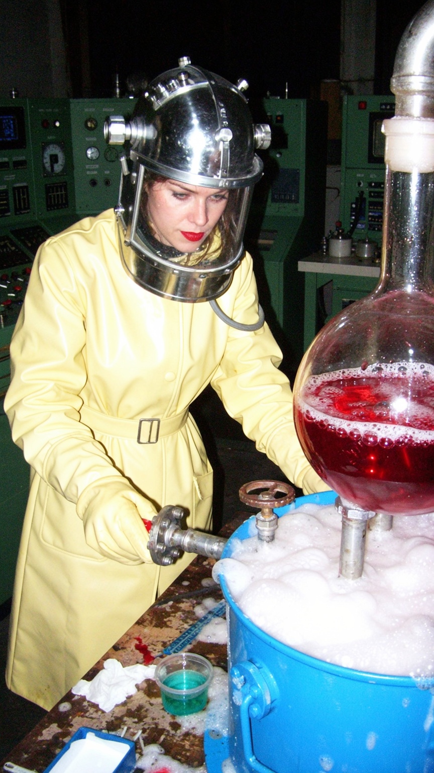 A candid snapshot of a woman in retro-futuristic laboratory protective gear operating an industrial chemical setup. She we...