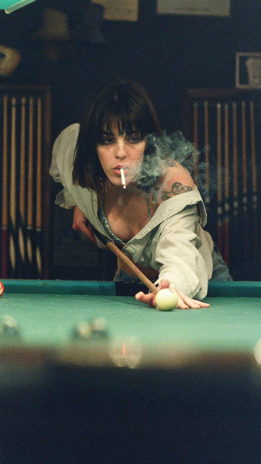 A super moody and cinematic shot of a cool girl with short dark hair and bangs,  leaning low over a pool table in a dimly...