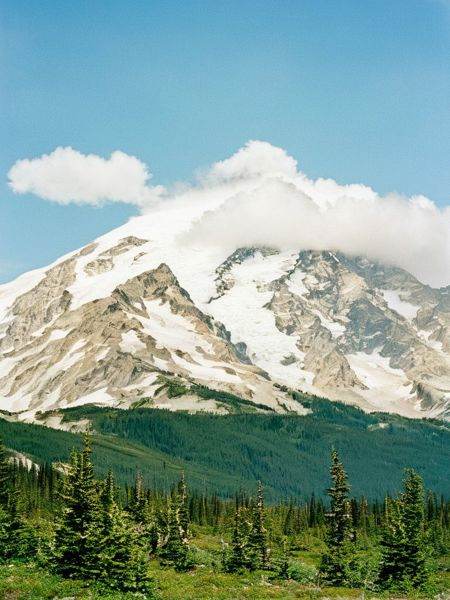 film photography style a snowy mountain range with a forest in the foreground,  fluffy clouds in the sky,  early morning o...