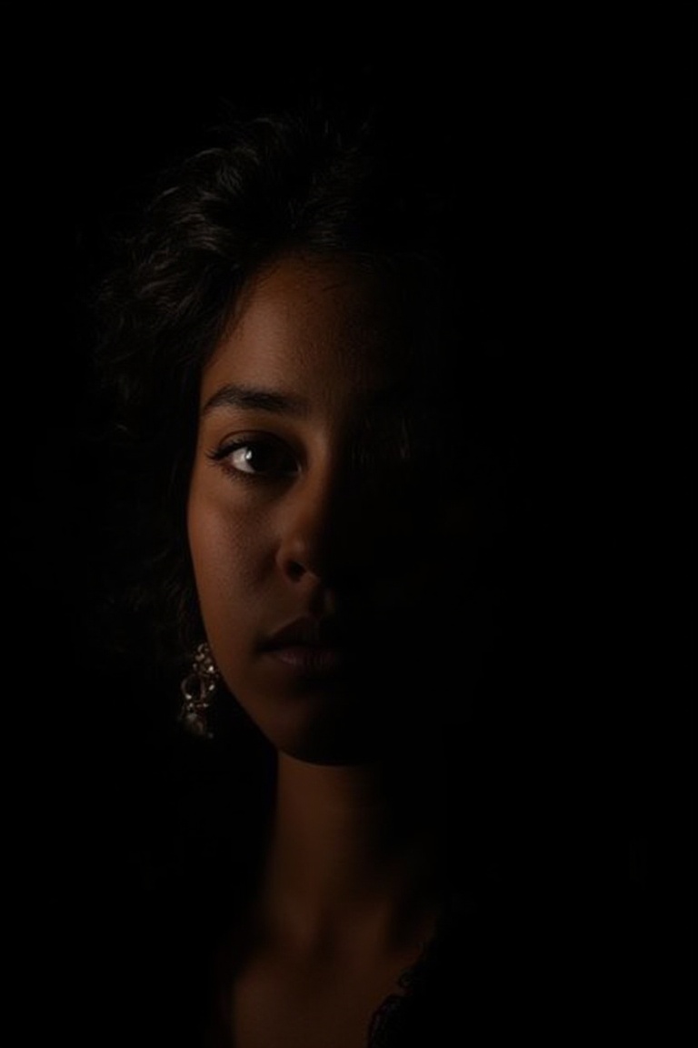 A Latina woman,  her curly hair spilling over her shoulders,  the only light softly tracing her face against the black bac...