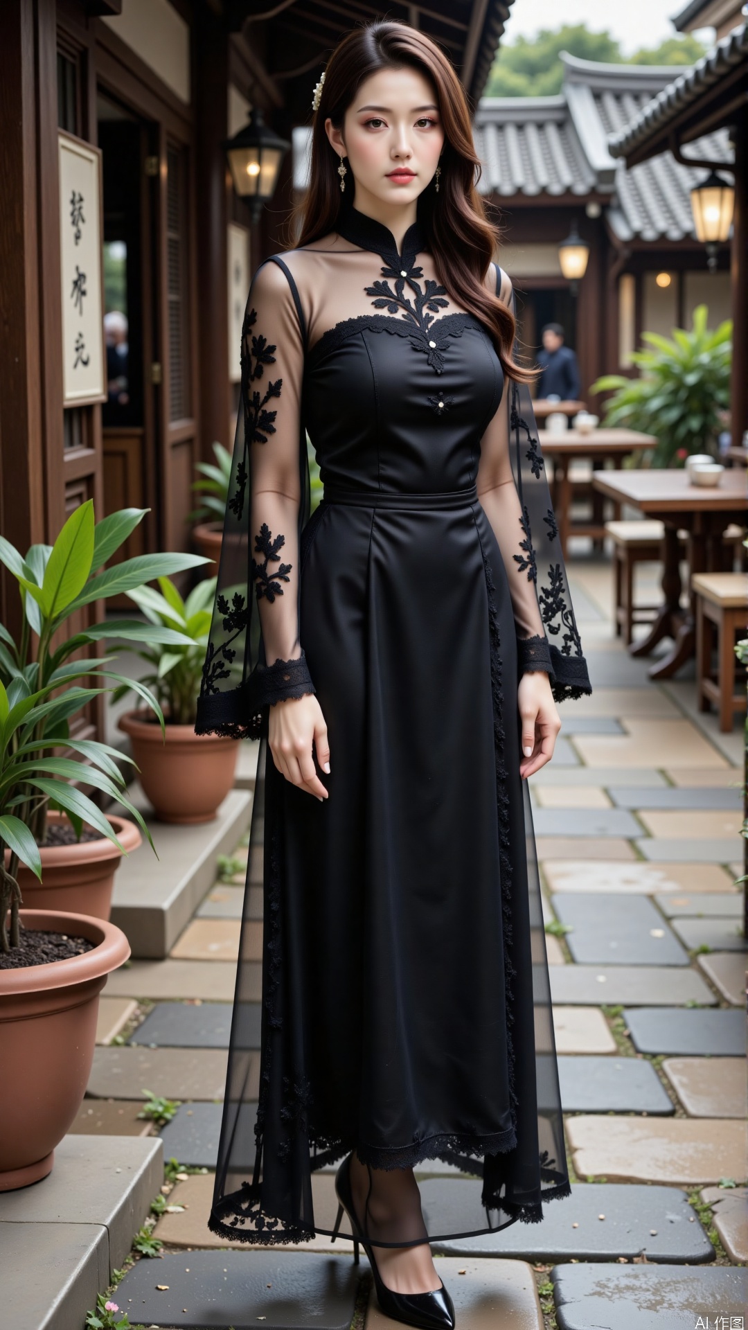 The image shows a woman standing outdoors in what appears to be a traditional or historical setting. She is wearing a black dress with sheer, embroidered sleeves that have a floral pattern. Her hair is long and wavy, styled with a hair accessory. The background includes a traditional building with wooden elements and potted plants. The ground is paved with stone tiles, and there are some tables and other objects visible in the background.
