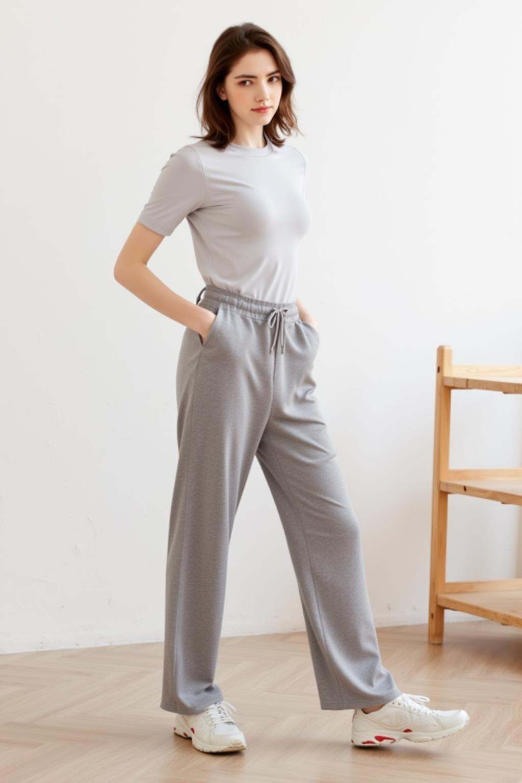 A woman is standing in a room with a white wall behind her. She is wearing a short sleeve shirt and gray pants. Her shoes are white and have red on the side of them. There is a wooden shelf on the floor next to the woman.