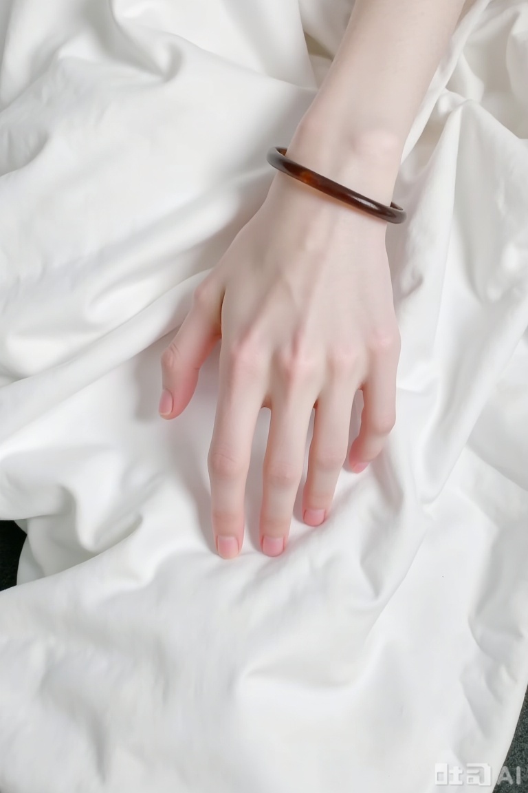 The image shows a close-up of a person's hand resting on a white fabric surface. The hand is positioned with fingers slightly curled, and the skin appears smooth and pale. The person is wearing a brown bracelet on their wrist. The background consists of a white, soft fabric, possibly a blanket or clothing, and a hint of a dark-colored surface at the bottom left corner. The overall setting is minimalistic and focused on the hand and the fabric.