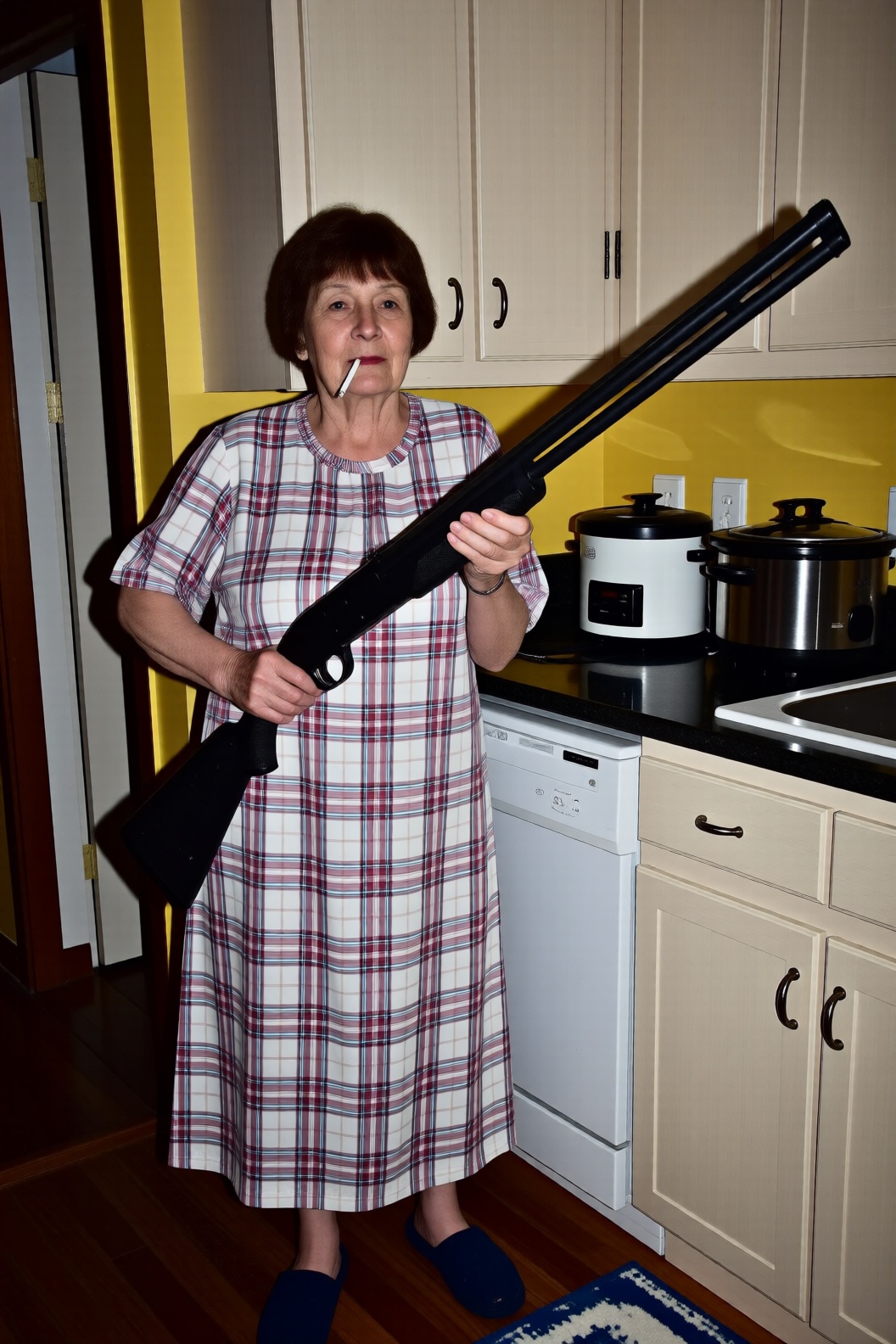 A full-body shot of an elderly woman standing in a domestic kitchen with bright yellow walls. She is wearing a loose,  lig...