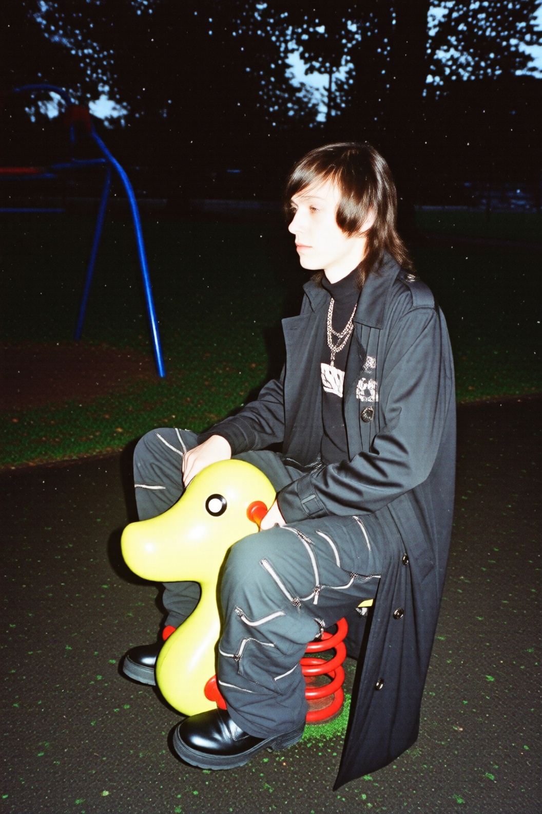 A young man dressed in full goth attire,  including a black trench coat and baggy pants with many zippers and chains,  sit...